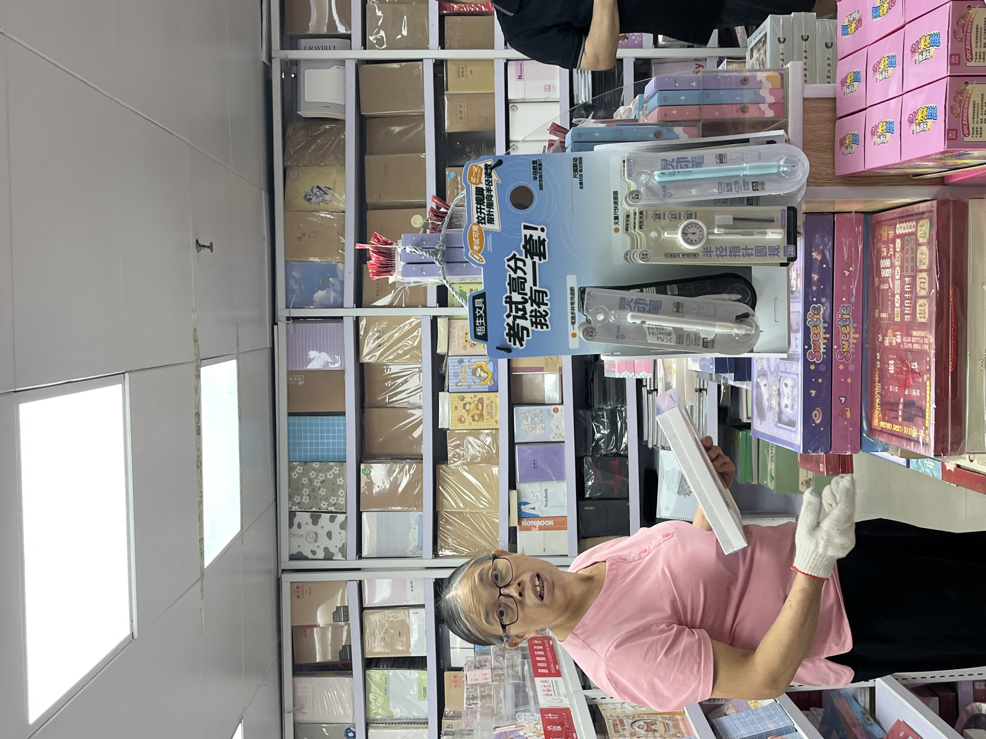 In a stationery store located in Liwan District, China, an elderly woman wearing a pink polo shirt and glasses stands in front of shelves stocked with notebooks, pens, and other school supplies. She holds a white box in her gloved hands, suggesting she is a customer or employee. The store appears well-organized with a wide selection of items displayed on white shelves. The lighting is bright, indicative of an indoor commercial setting. A display featuring stationery related to exams is prominently placed, showing various writing instruments and tools. The overall environment suggests a typical retail space dedicated to office and school supplies.