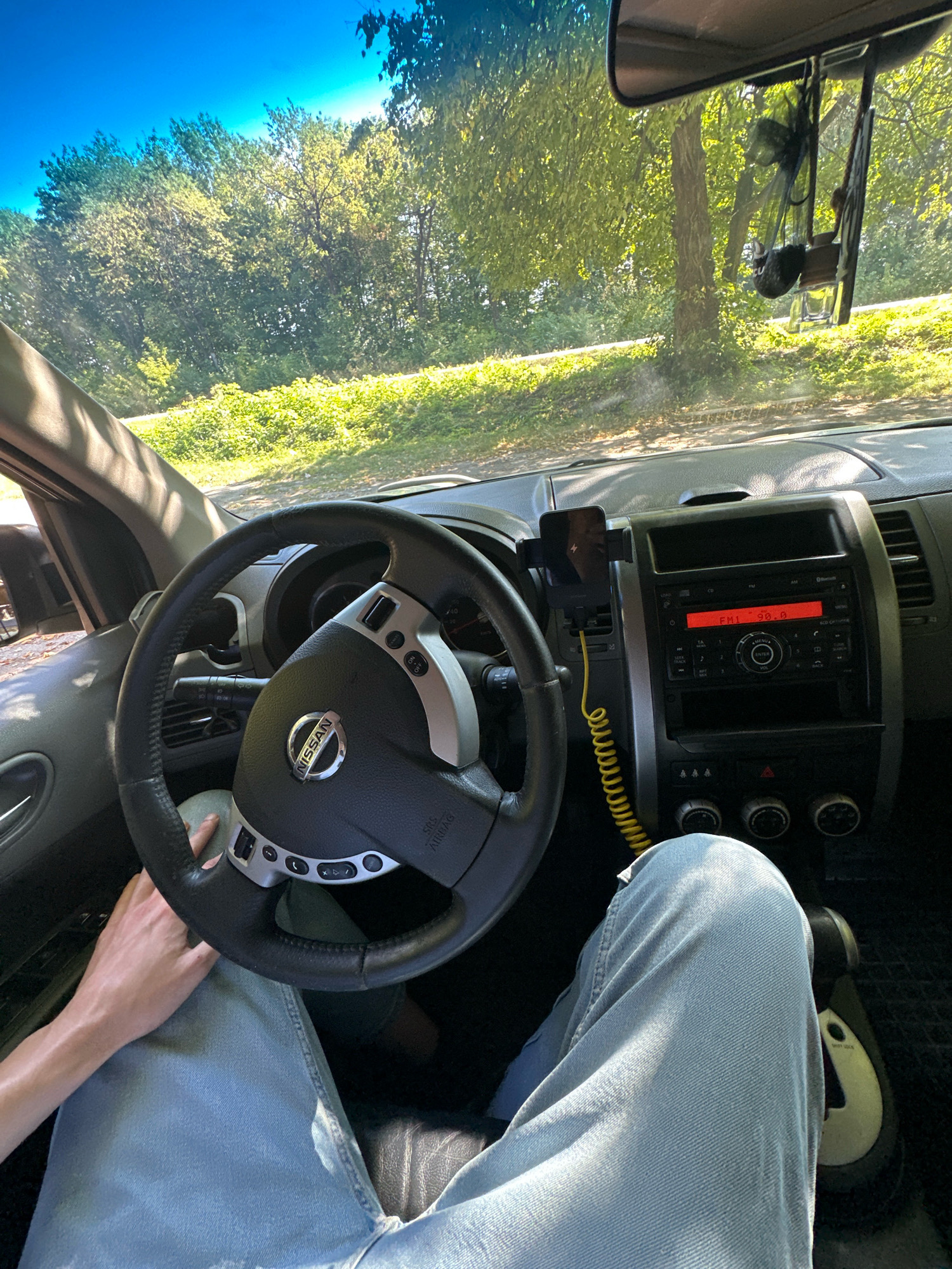 The image, taken from the driver's seat of a Nissan, offers a view of the car's interior and the sun-dappled trees and foliage outside. The driver's hands are visible, one resting on the steering wheel and the other on their lap. The car's radio is displaying "FM 90.0," suggesting it is tuned to a radio station. A yellow coiled cable connects a phone mount on the dashboard to a power source. The scene appears to be set in Shatava, Ukraine, based on the provided context. The overall impression is one of a peaceful drive through a wooded area on a sunny day.