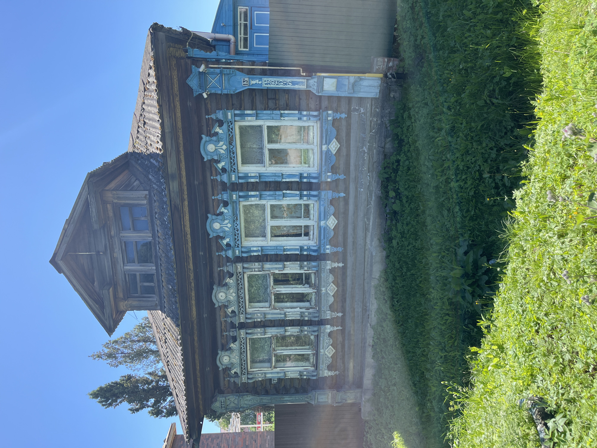 The image shows a weathered wooden house with ornate blue window frames and decorative carvings. The house is situated in a grassy area with some trees in the background. The sky is clear and blue. The architectural style suggests a traditional Russian rural dwelling. The house appears to be located in the village of Novo-Nikolskoye, Russia, indicated by the address number "135" on the wooden post next to the house. There are no people visible in the image. The overall impression is one of an old, rustic home in a sunny, outdoor setting.