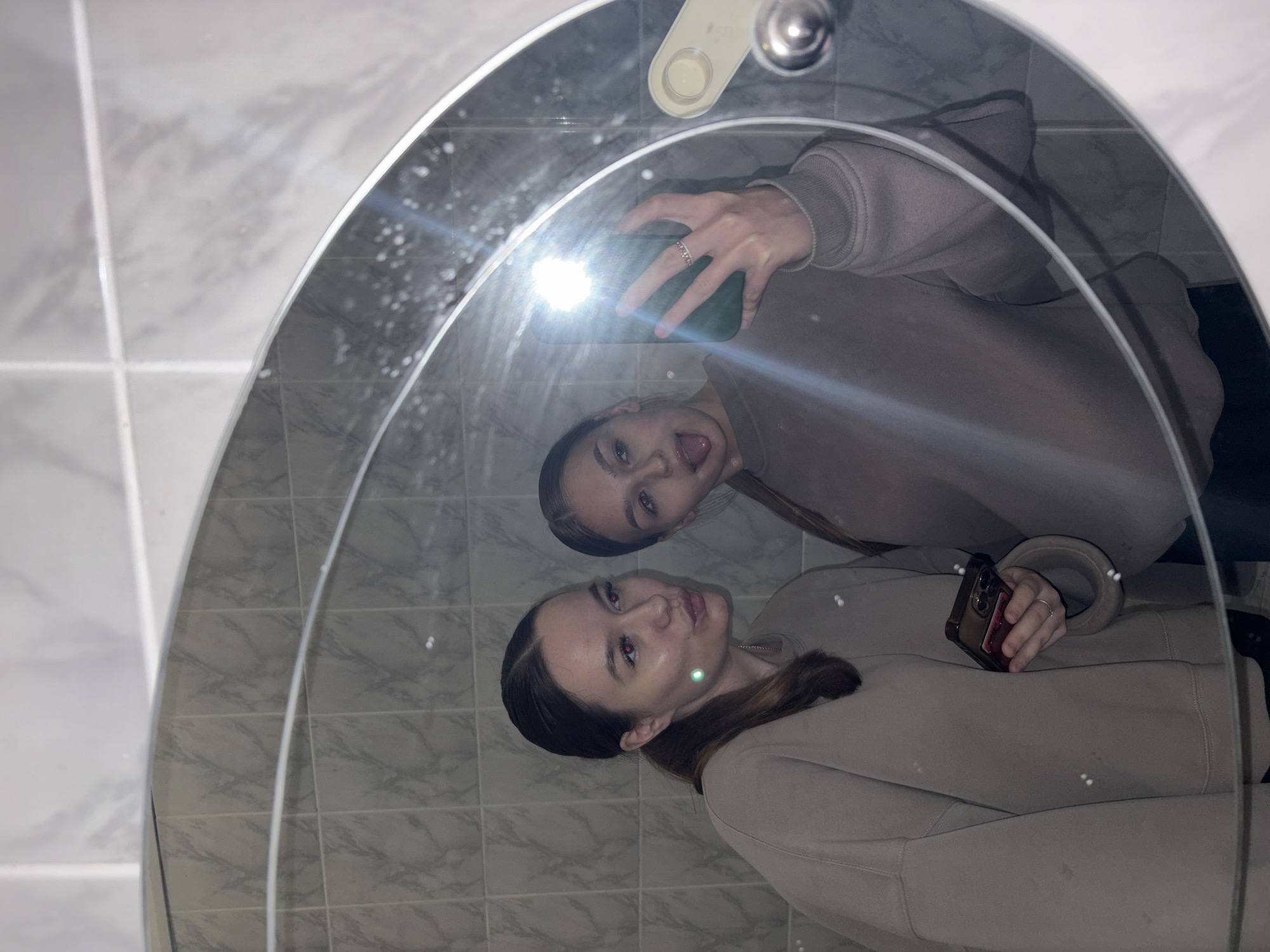 Two young women are taking a selfie in a bathroom mirror. The woman on the left is looking at the camera and puckering her lips. She has her hair pulled back in a ponytail and is wearing a beige sweatshirt. She is holding a phone in her left hand. The woman on the right is sticking her tongue out at the camera. She also has her hair pulled back and is wearing a beige sweatshirt. She is holding a phone in her right hand and has a ring on her finger. The background is a tiled wall and a mirror. The overall impression is informal and playful. The photo appears to have been taken in an indoor residential bathroom. There is no visible information in the image to suggest it is located in the city of Chaikovsky, Russia.
