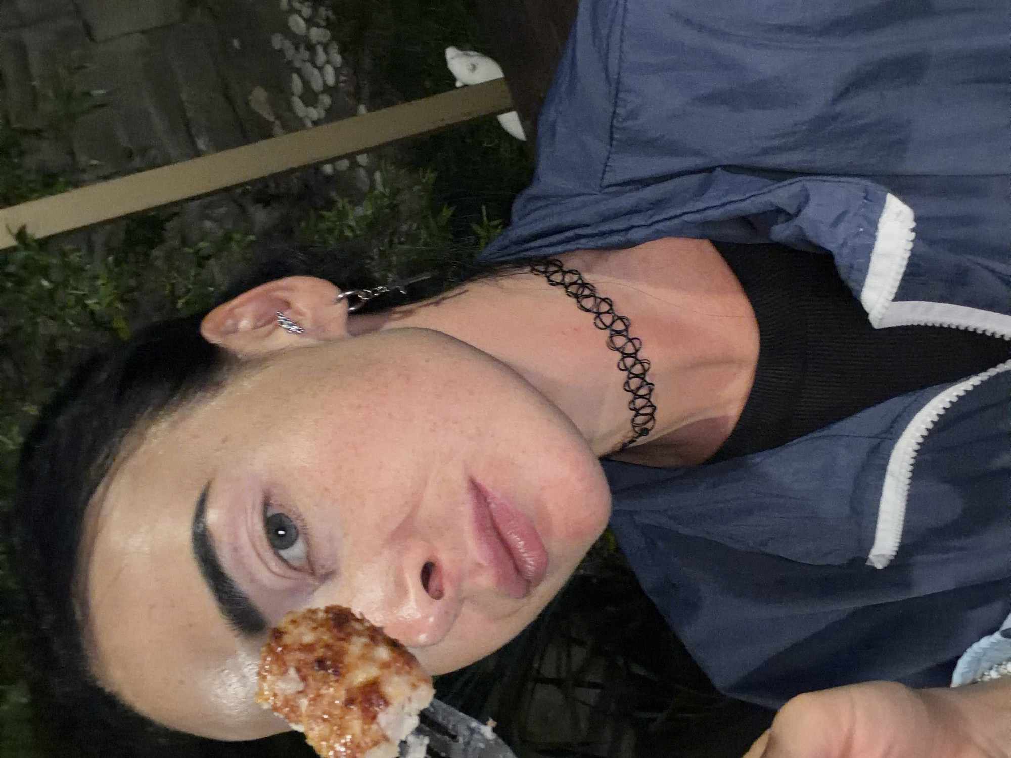 A woman is taking a selfie outdoors at night. She is holding a piece of food on a fork up to her eye. She is wearing a blue jacket over a black shirt, a black choker necklace, and earrings. There are trees and a white object in the background. The location is unknown.