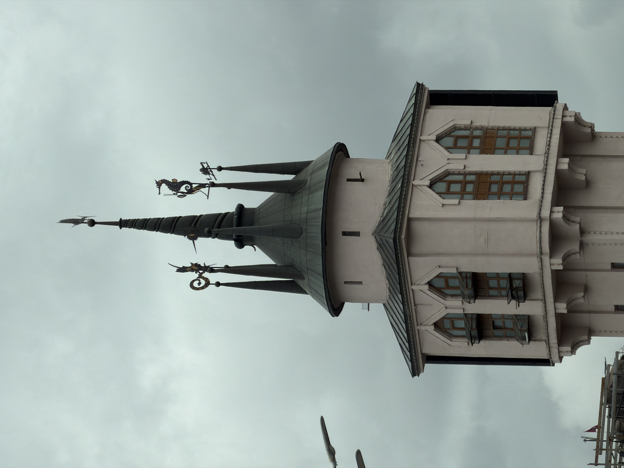 The image captures a distinctive architectural structure, likely a tower or a prominent section of a building, viewed from below against an overcast sky. The building features light-colored, possibly pinkish-beige, walls with rectangular windows set into recessed, geometric frames. Some windows appear to have open sashes.
The most striking feature is the complex, multi-spired roof structure atop the main building. The central spire is tall and pointed, topped with a finial. Several smaller, conical spires flank the main one, each adorned with ornate, dark-colored metallic weathervane-like decorations. These decorations depict fantastical creatures, including a dragon-like figure with a coiled tail and possibly another creature resembling a griffin or seahorse. The roof itself is dark, appearing to be made of metal or slate, with visible ridges and architectural detailing. The overall aesthetic suggests a historic, possibly Art Nouveau or revivalist architectural style.
The sky is uniformly cloudy and gray, indicating an overcast day. No people or specific activities are visible, and there is no discernible text on the building. The perspective is tilted, giving a dynamic view of the architecture reaching towards the sky. The location context provided is Moscow, Russia.