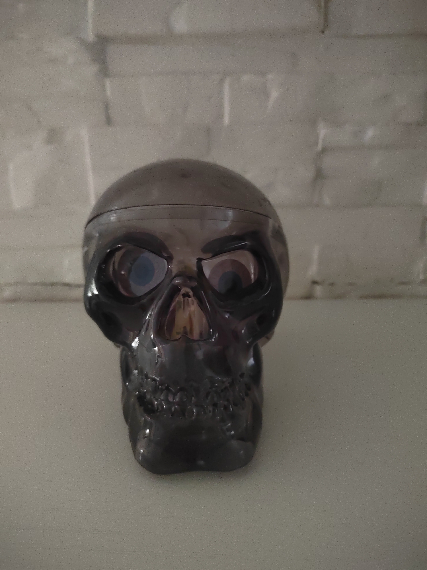 A close-up, eye-level shot shows a dark gray, transparent skull-shaped object with exaggerated eye sockets and a detailed jawline. The skull is positioned centrally on a light-colored, flat surface, likely a shelf or table. Behind the object, a textured wall, appearing to be made of light-colored bricks or textured plaster, forms the background. The lighting is even, with soft shadows. The image does not contain any discernible elements that would suggest its location within the city of Porto Recanati, Italy.