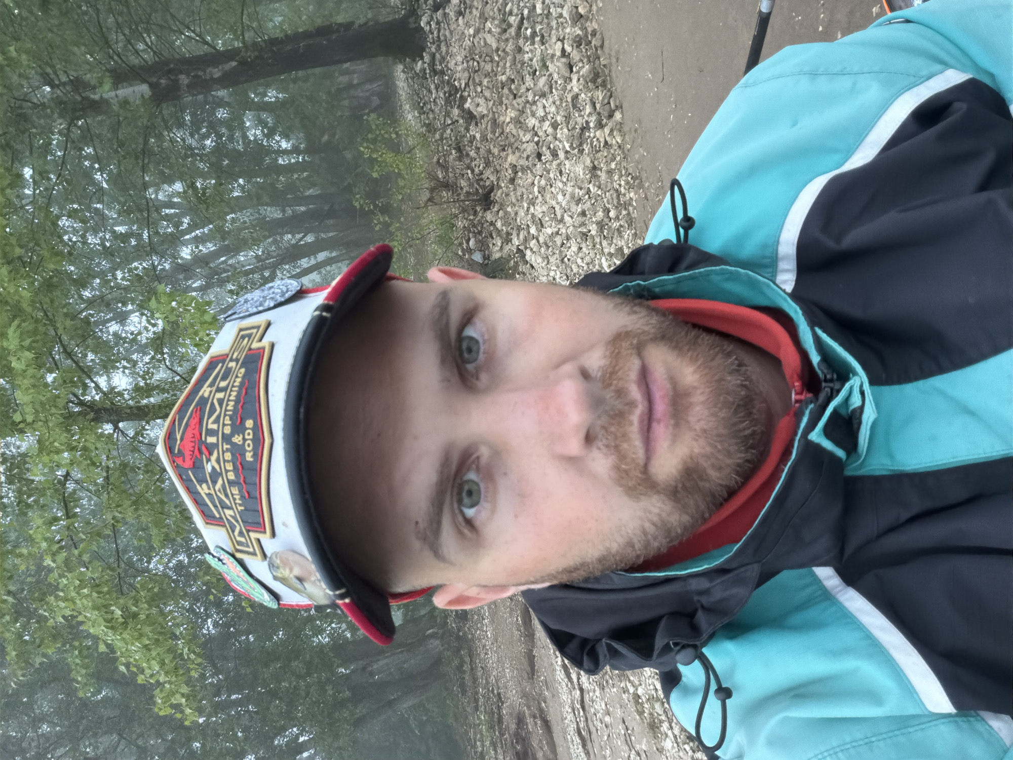 The image is a selfie-style, medium close-up portrait of a man, likely taken outdoors in Chaikovsky, Russia. The man looks directly into the camera with a neutral expression. He has light-colored eyes and a short, well-maintained beard.
He is wearing a white baseball cap with a red brim and trim. The cap prominently displays a logo that reads "MAXIMUS" in gold and red, with the slogan "THE BEST SPINNING & RODS" printed beneath it, suggesting a connection to fishing gear. Several pins or patches, including one shaped like a green fish lure, are attached to the cap. The man is also wearing an outdoor jacket, which is primarily light blue/turquoise and black, featuring a white stripe across the chest and arm. A red inner layer or collar is visible beneath his jacket.
The background suggests an overcast or foggy day. Behind the man, a dense treeline with green foliage is visible, partially obscured by mist or haze, indicating damp weather conditions. To his right, a textured ground of gravel or small stones is apparent, possibly a path, road, or riverbank. The overall scene conveys an outdoor, natural setting. The image is rotated 90 degrees counter-clockwise.