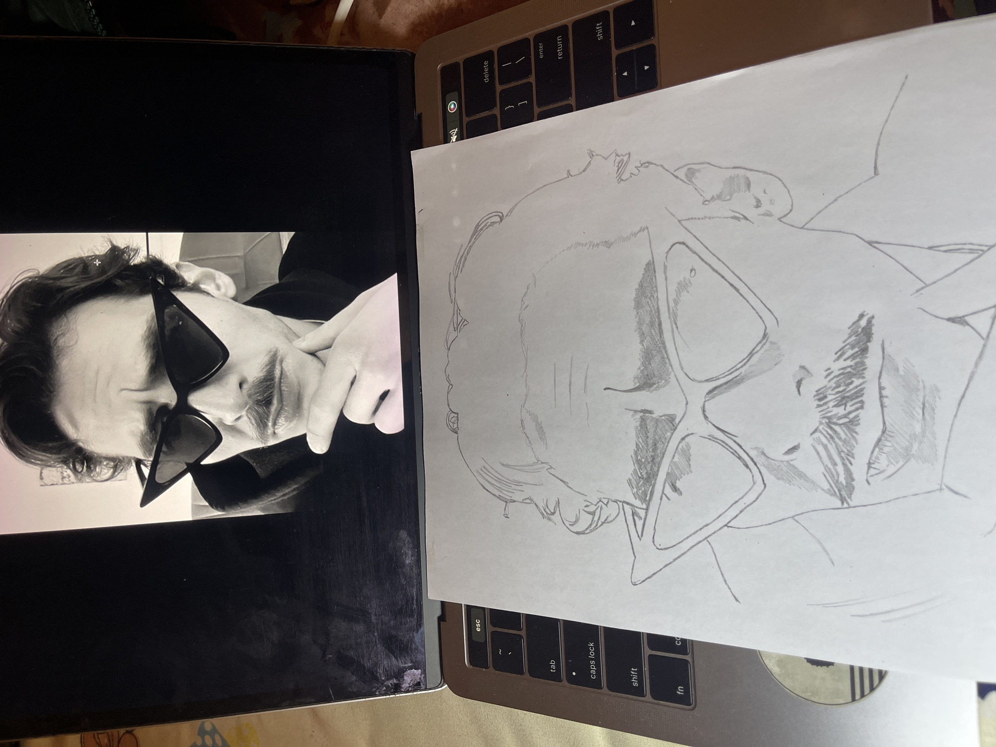A color photograph shows two laptops and a drawing of a man. The laptop on the left displays a black and white photograph of a man with dark hair, wearing angular sunglasses, and holding his fingers near his mouth. The laptop on the right has a sheet of white paper on top, displaying a pencil drawing of the same man. The drawing replicates the pose and features of the man in the photograph, including his sunglasses and facial hair. Both laptops are resting on an indistinct surface. There is no information about Jos, Nigeria, or any details of a city.