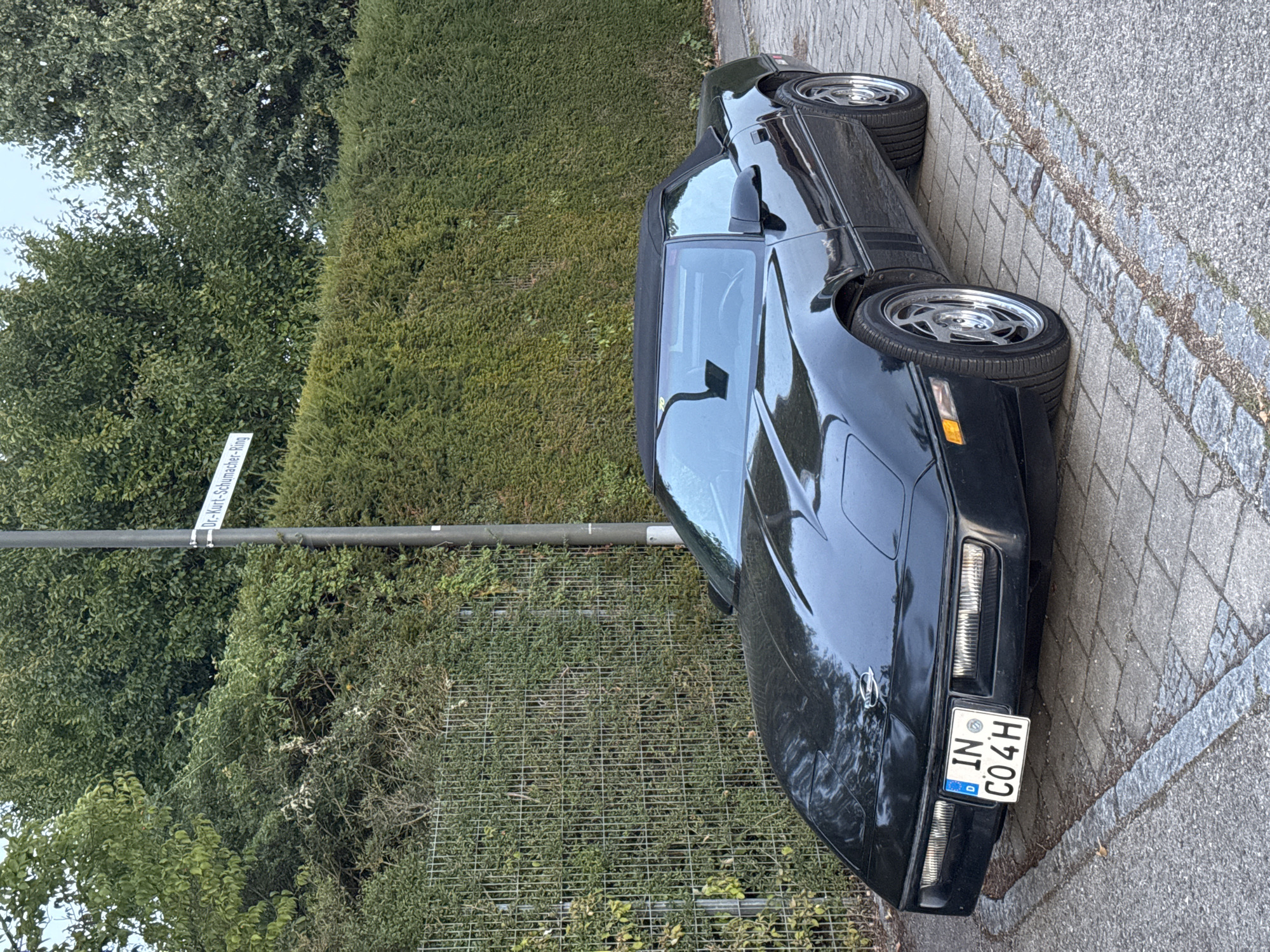 In the city of Wettstetten, Germany, a black convertible Chevrolet Corvette C4 is parked on a paved street with a cobblestone border. The car is facing slightly towards the right of the frame. In the background, a metal fence is visible, partially covered by greenery. Beyond the fence, a tall, dense hedge forms a boundary. A street sign reading "Dr.-Kurt-Schumacher-Ring" is mounted on a pole to the left of the car. The sky is overcast.