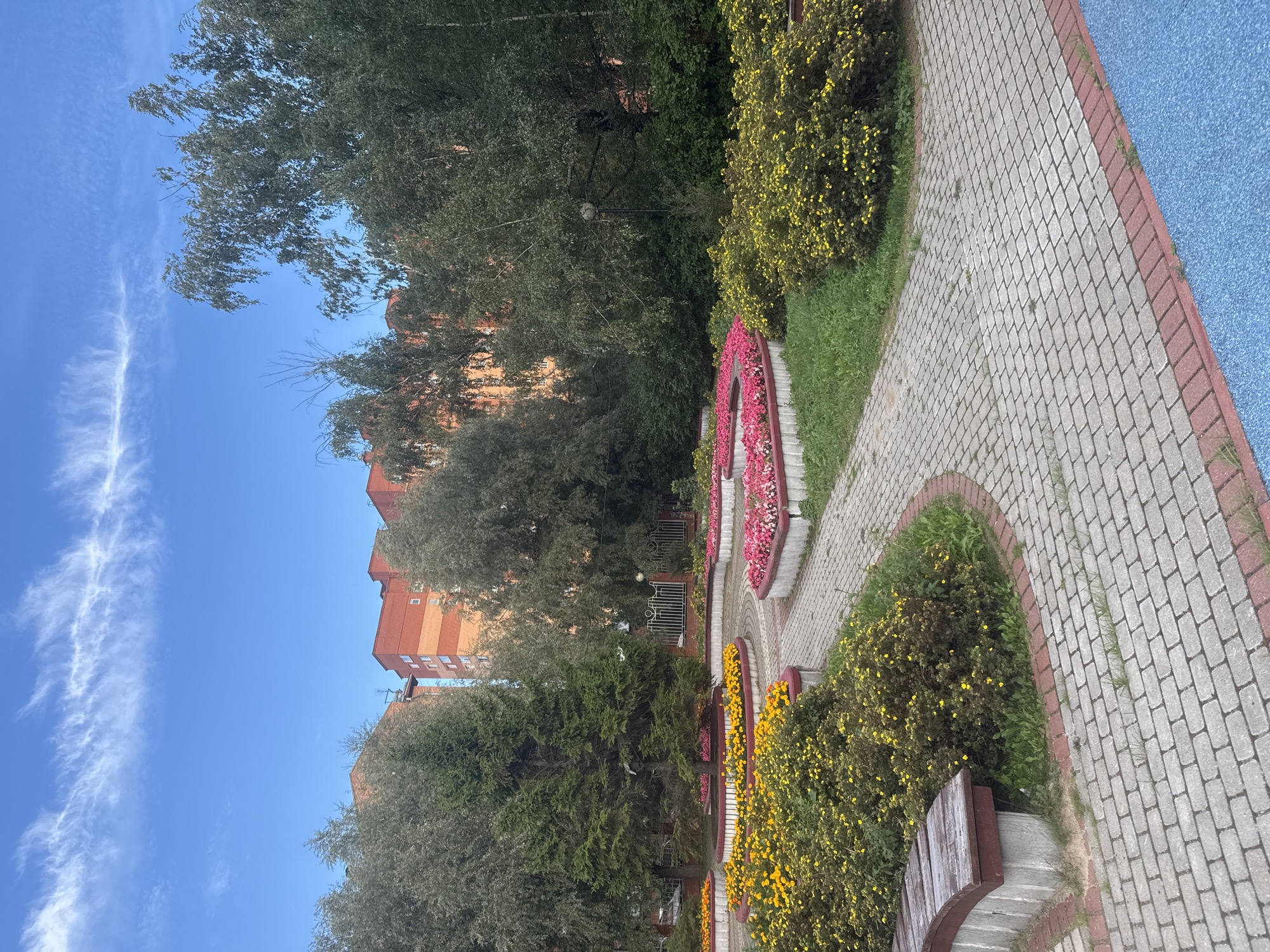 The image depicts a courtyard in the city of Sovkhoz imeni Lenina, Russia. The foreground features a paved walkway made of grey interlocking bricks with a red brick border. To the left, a wooden bench with a concrete base is situated next to a circular flower bed filled with yellow flowers and green shrubs. Adjacent to this, another curved flower bed contains vibrant pink flowers. Further back, more flower beds with pink and yellow blooms are visible, interspersed with grass and trees. In the background, several multi-story residential buildings, predominantly orange and red brick, are visible behind a line of trees. The sky is a clear blue with a few wispy white clouds. The scene appears to be an outdoor public space designed for recreation or aesthetic appeal.