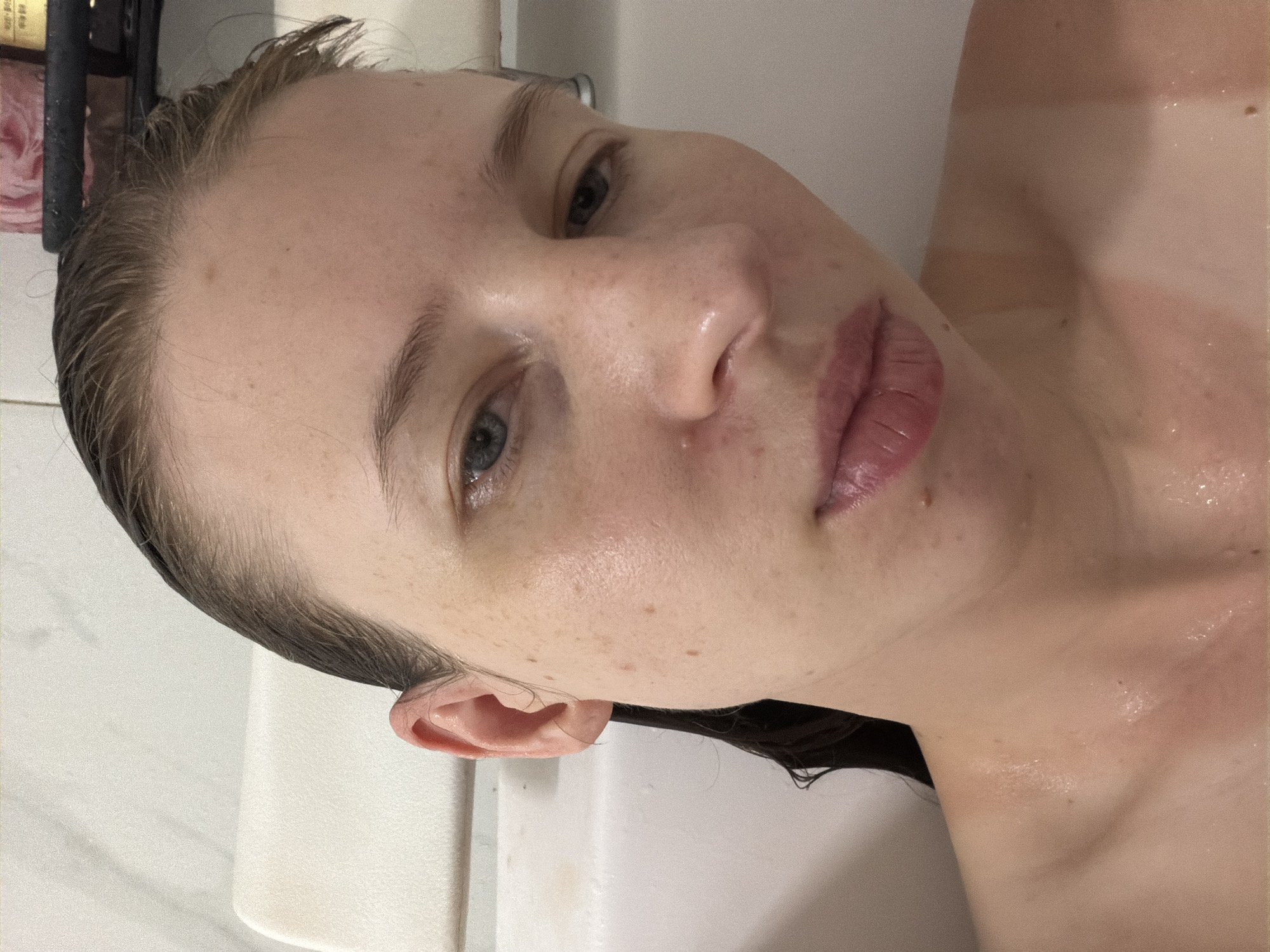 A close-up, overhead, selfie-style shot shows a person's face, ears, neck, and upper chest, positioned in a bathtub in a dimly lit room. The person, whose face is moist and appears to be wearing no makeup, has light skin with visible freckles and some small blemishes. Their hair is wet and slicked back, revealing the curve of their ears and the hairline. The backdrop consists of a white bathtub and a dark shelf with a pink bottle containing a liquid. The lighting casts a warm glow on the person's skin, highlighting droplets of water on their face, neck, and chest, suggesting they are either in or just finished a bath. The image does not provide enough information to determine the location within Dongducheon-si, South Korea.