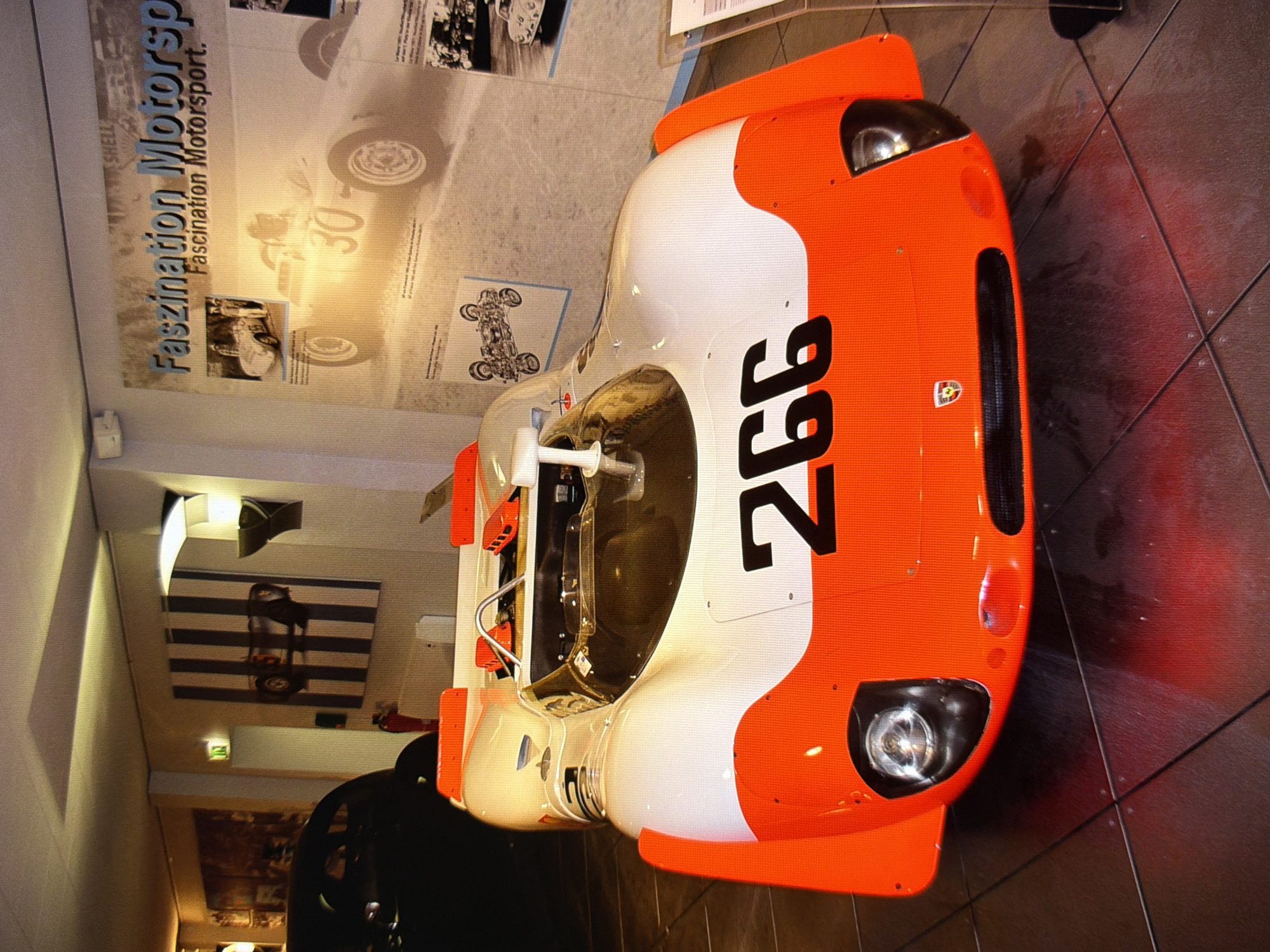 The image depicts a white and orange race car with the number 266 prominently displayed on its front. The car is positioned in what appears to be a museum or exhibition space, with tiled flooring and a wall displaying posters and artwork related to motorsports. One poster features the text "Faszination Motorsport." A black vehicle is partially visible in the background to the left of the race car. The lighting suggests an indoor environment, possibly a dimly lit exhibition hall. There are no people clearly visible in the foreground or interacting with the car. The scene is static and appears to be a capture of an exhibit. The location is identified as Kyiv, Ukraine.