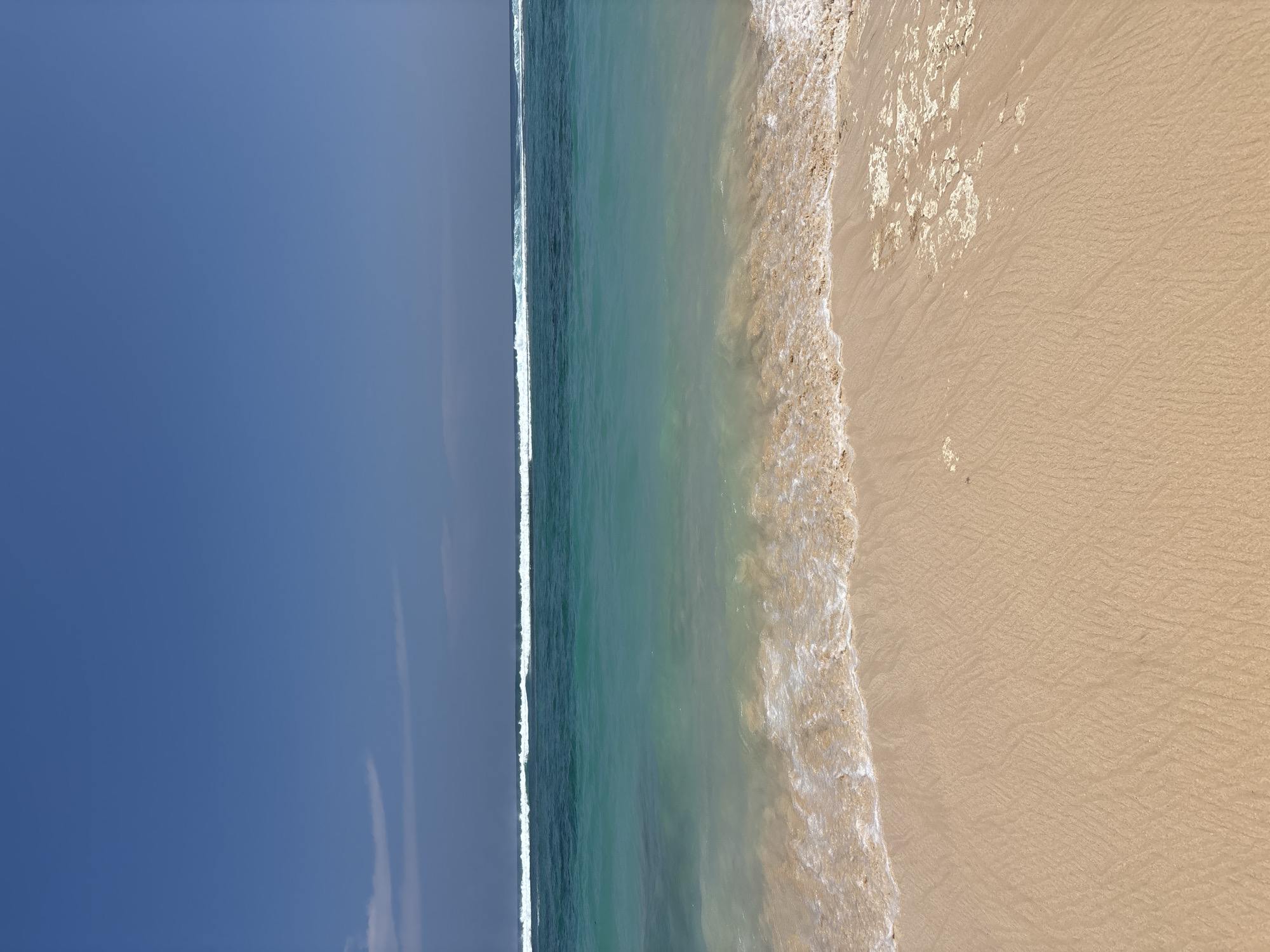 The image displays a clear daytime view of a beach and ocean in Ungasan, Indonesia. The upper approximately one-third of the frame consists of a deep blue sky with very faint, elongated white cloud formations present in the upper left area. Below the sky, the ocean is visible, horizontally stratified into distinct color zones. The farthest ocean segment presents a line of white breaking waves against a darker blue-green water mass. This transitions into a wide band of clear turquoise water, which then lightens to a translucent green-blue as it meets the shoreline. The lower half of the image is occupied by light tan, fine-grained sand. The wet sand exhibits intricate ripple patterns consistent with receding wave action. In the lower right foreground, irregular patches of light-colored, textured material, appearing as dried coral fragments or mineral deposits, are scattered across the sand. No people, animals, man-made objects, actions, or interactions are depicted within the scene.