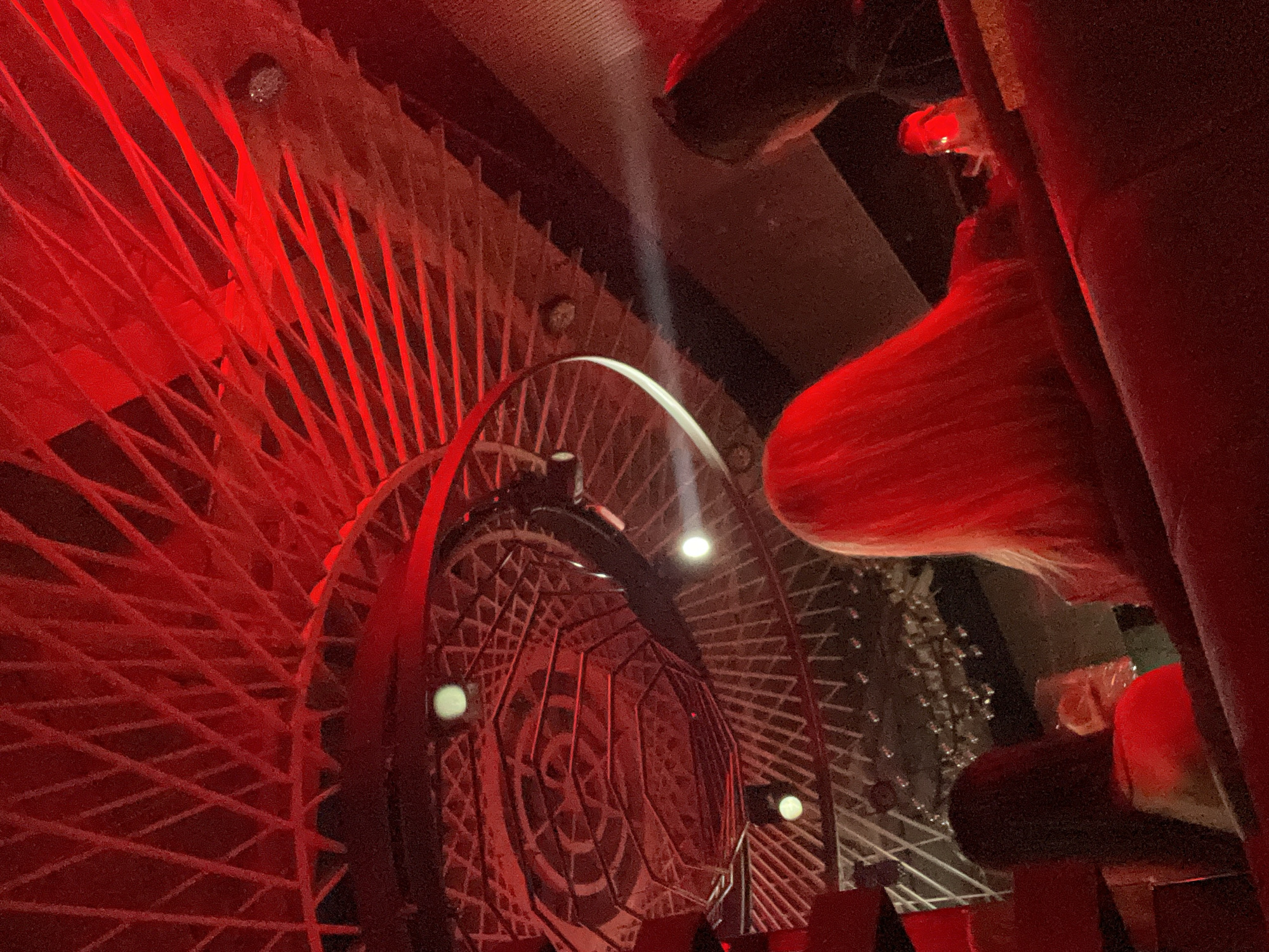 The image depicts an indoor entertainment or dining venue, possibly a bar or club, in an unknown city. The dominant lighting is a deep red, cast throughout the space, creating a vibrant and intense atmosphere. The ceiling features multiple intricate layers of architectural design. The uppermost layer consists of a criss-crossing pattern of narrow beams or slats. Below this, a large circular structure showcases an elaborate geometric pattern, possibly composed of concentric octagons and repeating motifs, illuminated in red. Several bright, circular white spotlights are integrated into these ceiling structures.
In the foreground, the back of red upholstered seating, likely a booth or bench, is visible. At least three individuals are partially visible from behind, seated and facing away from the camera. On the left, a person with dark hair is holding a transparent glass. In the center-right, a person with long, blonde hair is prominent, also facing away. To the far right, another person with dark hair is visible, largely obscured. A distinct, narrow beam of white light cuts horizontally through the red ambiance on the right side of the frame, originating from an unseen source and illuminating the space in front of the blonde-haired individual. On the table in front of the blonde-haired person, a small, glowing red object, possibly a drink or a decorative light, is present. No explicit interactions between the individuals are discernible.