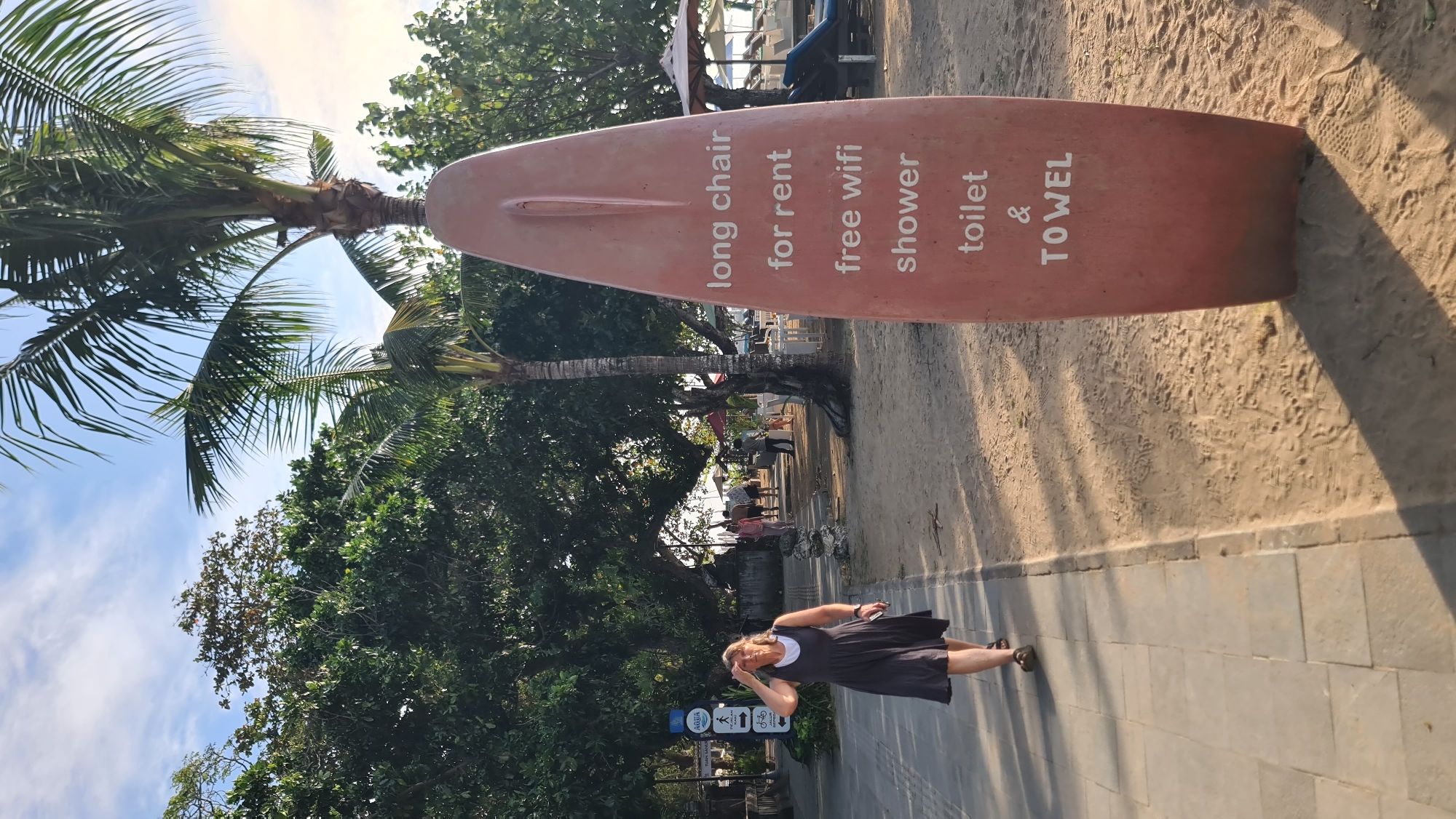 The image shows a woman walking on a paved path next to a sandy beach in Denpasar, Indonesia. She is wearing a dark dress and has her hand on her forehead, shielding her eyes from the sun. Behind her, several palm trees and lush green foliage line the path. In the foreground, a large, red surfboard-shaped sign is planted in the sand, with white lettering listing amenities: "long chair for rent", "free wifi", "shower", "toilet & TOWEL". In the background, beyond the trees, beach chairs and umbrellas are visible, suggesting a resort or public beach area. The sky is partly cloudy, and the lighting indicates it is either early morning or late afternoon.