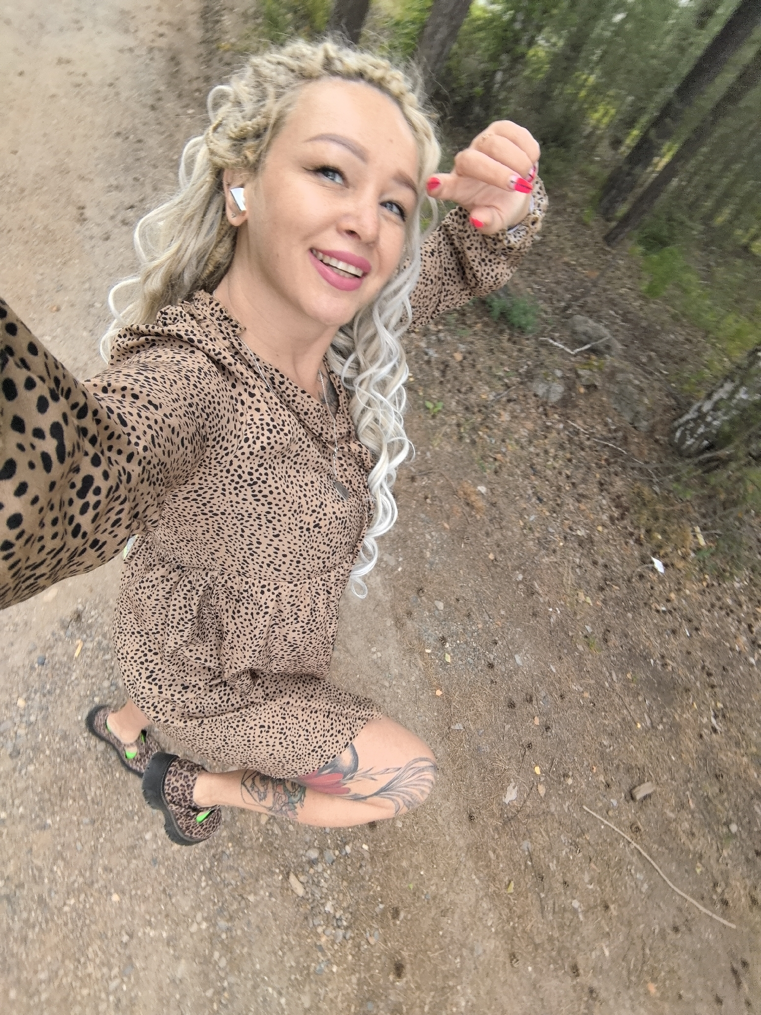 A woman is captured in a selfie outdoors, kneeling on a dirt path with a backdrop of trees. She has blonde, braided hair and is smiling brightly, her makeup including pink lipstick and winged eyeliner. She's wearing a leopard-print dress with a ruffle detail at the neckline and matching leopard-print shoes with green accents. Her left hand, with bright pink nail polish, is raised towards the camera, with a ring visible on one finger. Tattoos are visible on her left leg. The setting appears to be a wooded area, with pine cones and dry leaves scattered on the ground. The lighting suggests it's daytime, likely overcast given the diffused light and the lack of strong shadows. The woman's expression and the selfie pose convey a sense of joy and engagement.