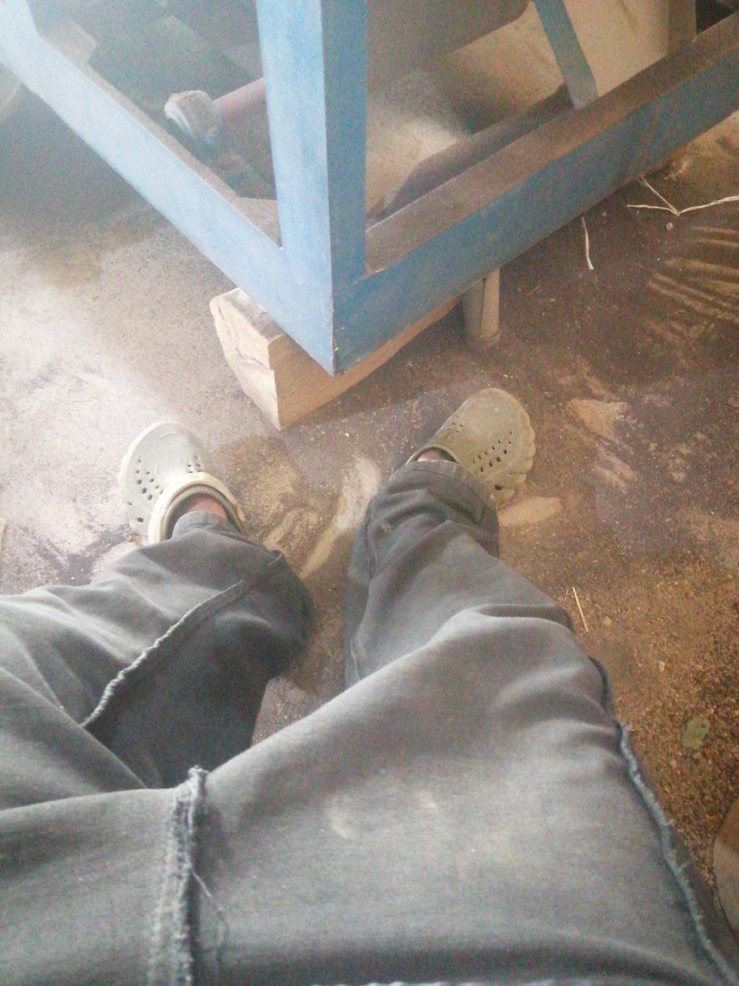 The image shows a person's legs and feet, wearing grey trousers and light-colored clogs, sitting on the ground. The ground appears to be dusty or sandy and there are some scattered wood fragments. In the background, a blue metal structure, possibly part of a machine or equipment, is visible. There are no specific indicators to determine the exact location within Jos, Nigeria, but the scene suggests an outdoor or workshop environment. The lighting is bright, and the image quality is somewhat low, making it difficult to discern finer details.