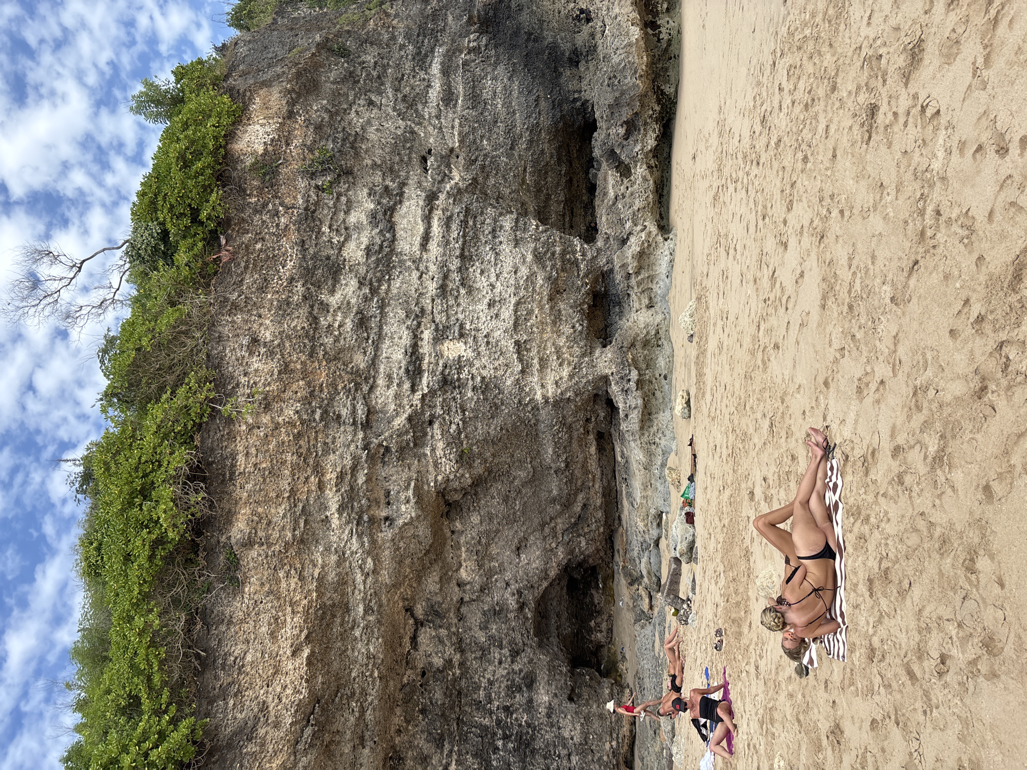 A group of people are sunbathing on a sandy beach in Kuta Selatan, Indonesia. The beach is backed by a large, rocky cliff face with some vegetation growing at the top. The sky is partly cloudy with blue skies. In the foreground, two women are lying on towels, facing each other. One woman is wearing a black bikini, and the other is wearing a black bikini top and bottoms. Further up the beach, closer to the cliff, several other people are seen sunbathing and walking. One person is standing near the cliff face, wearing a red swimsuit and a straw hat. The sand on the beach is light brown and has footprints scattered across it. The rocky cliff has several indentations and caves.