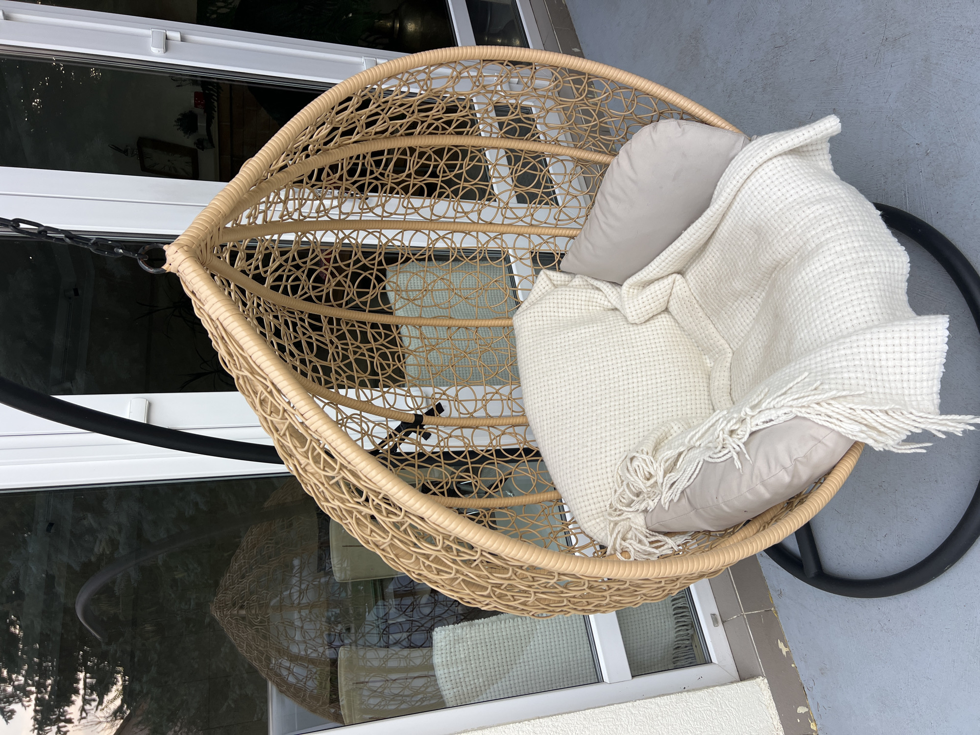 A wicker hanging egg chair with beige cushions and a white waffle-knit throw blanket is positioned on a grey tiled patio in front of a large glass sliding door. The chair is suspended by a black metal chain from a black metal stand. Reflections of trees and interior elements, including a clock and plants, are visible in the glass door. The scene is located in Fontanka, Ukraine.