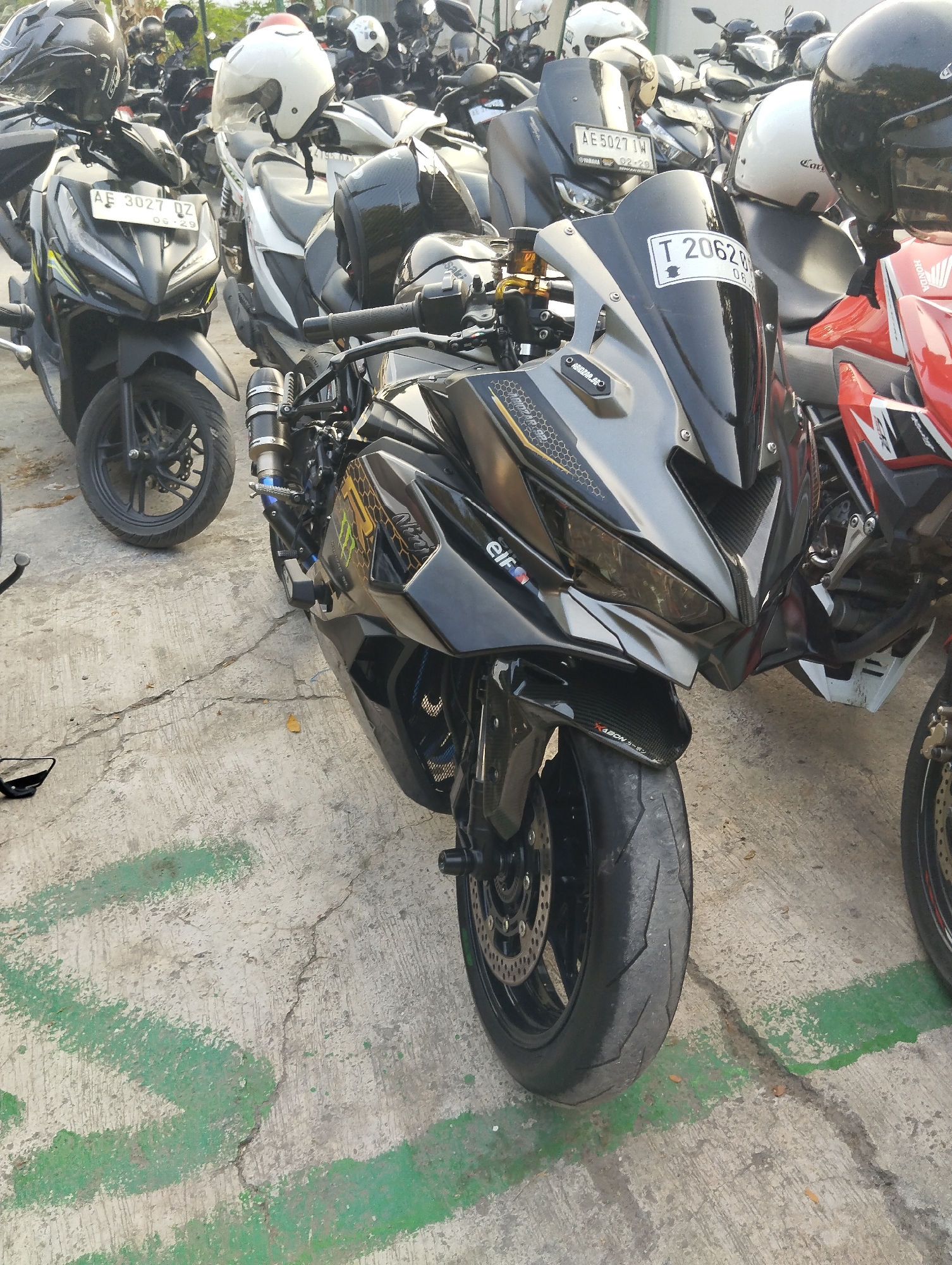 A black sport motorcycle is parked on a concrete ground with green markings. It is equipped with a black windscreen, black front fairing, and a gold-colored brake reservoir. A license plate with the text "T 2062 BG" and "08-26" is attached to the front. The motorcycle is partially obscured by another red sport motorcycle on its right. In the background, several other motorcycles and helmets are visible, suggesting a parking lot. The lighting and shadows indicate it is daytime.