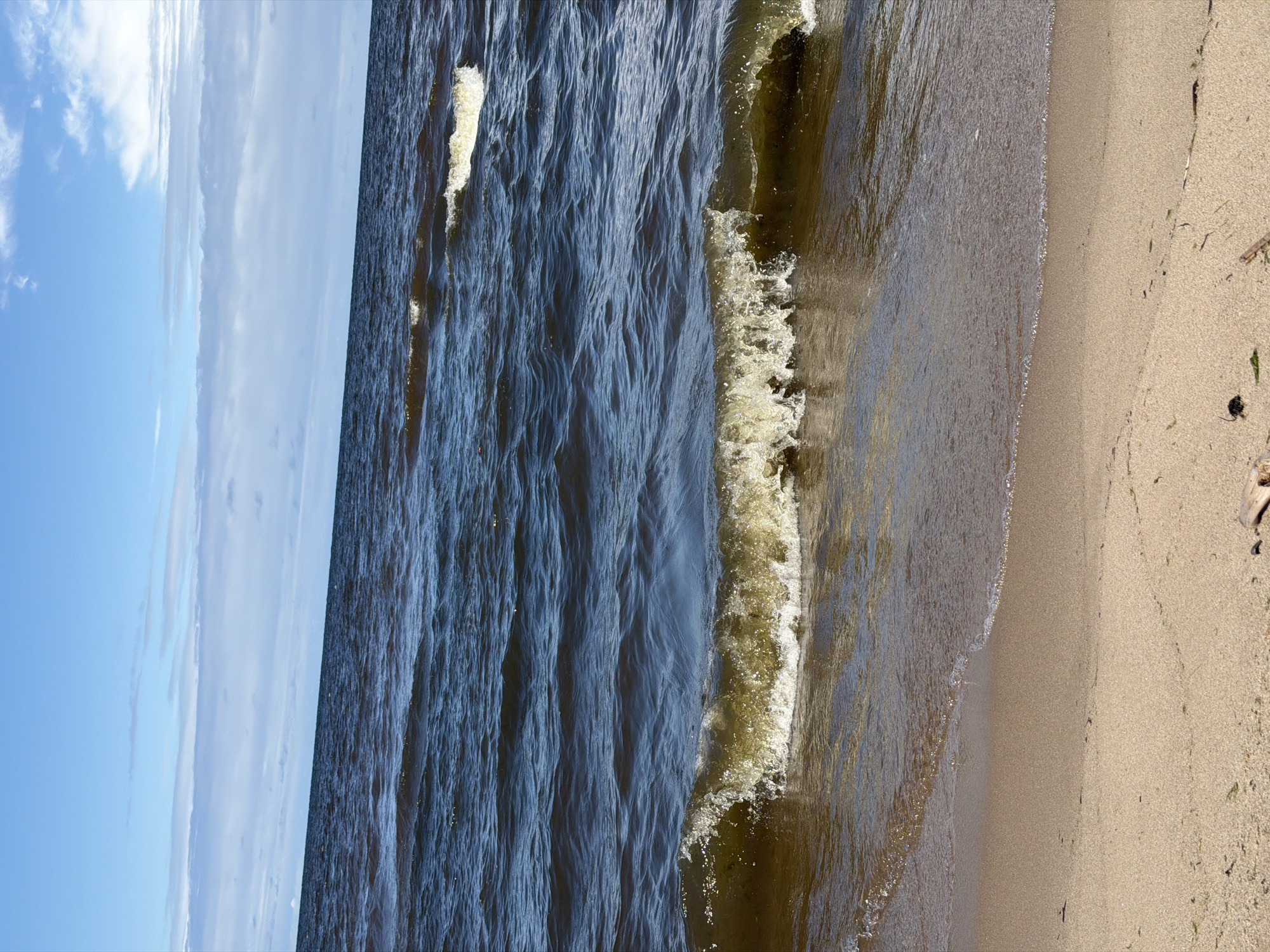 A wide-angle shot shows a body of water with waves breaking on a sandy shore. The water is dark and choppy, with some whitecaps visible. The sky is blue with scattered white clouds. The beach is light brown and appears to be composed of fine sand. Some small pieces of driftwood and seaweed are scattered on the beach. The scene appears to be a natural landscape, possibly a lake or a sea. This image was taken in Gauja, Latvia.