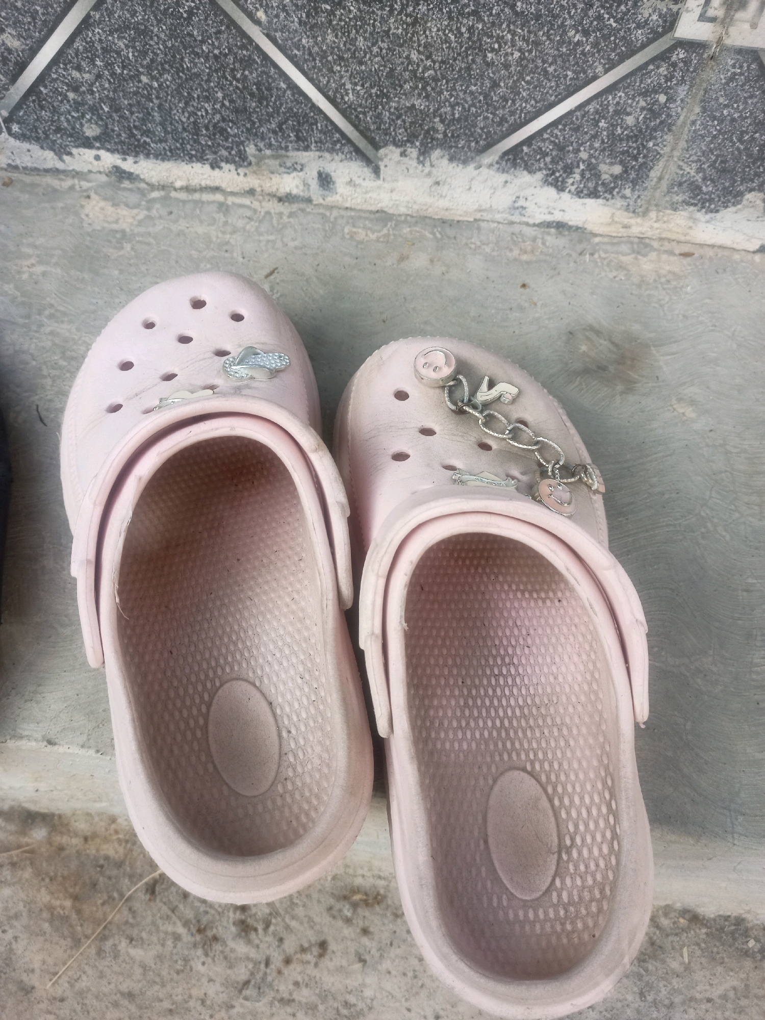 A pair of pale pink Crocs-style clogs are placed side-by-side on a concrete surface in front of a textured grey wall with silver accents. The clogs are decorated with charms, including a small silver flip-flop on the left clog and a chain with smiley face charms and a high-heeled shoe charm on the right clog. The interior of the clogs shows some signs of wear and dirt. The environment appears to be an outdoor space, possibly a porch or walkway, in Karangtalun, Indonesia.