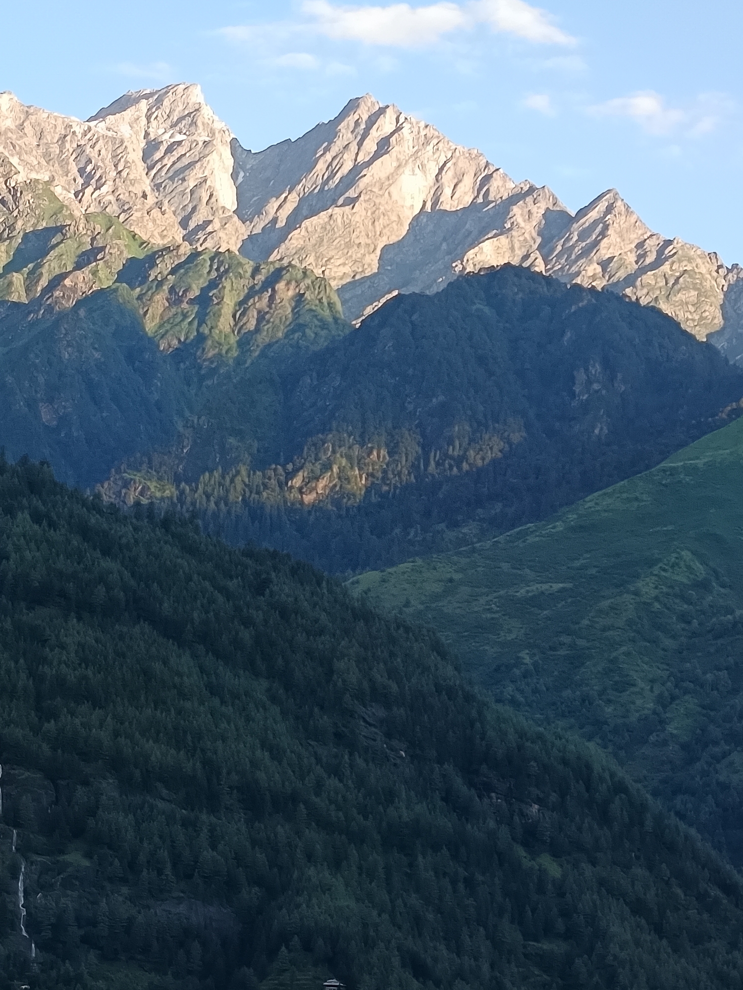 The image depicts a panoramic view of a mountainous landscape, likely in the vicinity of Vashisht, India, given the context. The scene is dominated by several layers of mountains, receding into the distance. The uppermost layer consists of rugged, snow-capped peaks bathed in the warm light of what appears to be either sunrise or sunset, casting sharp shadows that accentuate the jagged contours of the rocks. Below these peaks, successive layers of forested mountains descend. The mid-ground mountains are densely covered with dark green coniferous trees, with some patches of lighter green vegetation visible, possibly due to sunlight filtering through. The foreground features a steep mountainside also covered in a thick carpet of dark green trees. In the lower left corner of the image, a thin white stream of water, likely a waterfall, cascades down the mountainside. The sky above is a clear blue, with a few wispy white clouds present. No people or animals are visible in the image. The environment is natural and remote, characterized by rugged terrain and dense forestation.