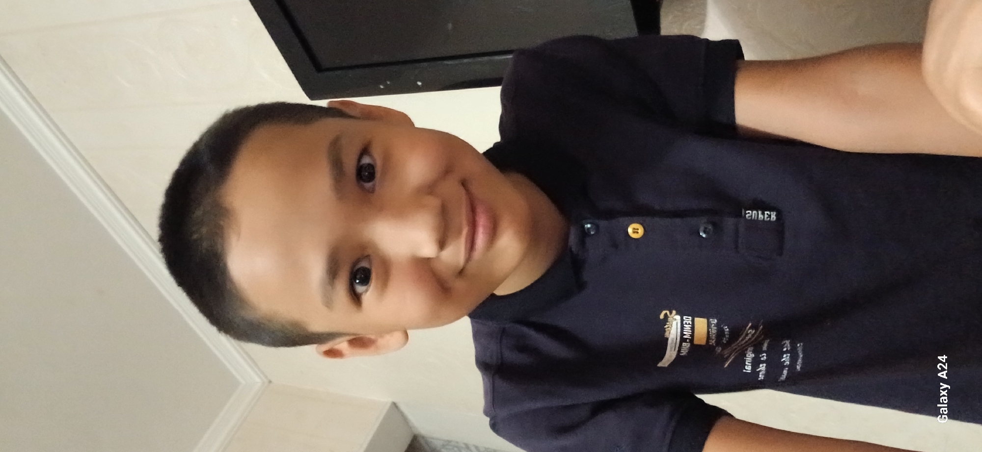A young boy with short, dark hair and dark eyes is looking at the camera and smiling. He is wearing a dark blue polo shirt with a logo on the left side of the chest. The shirt has two buttons at the collar, one of which is yellow. He is in an indoor setting with a plain, light-colored wall behind him and a dark object, possibly a television or monitor, to his right. The overall lighting is neutral. The "Galaxy A24" watermark is visible in the lower left corner of the image. The image appears to be a selfie taken indoors, with no indication of its specific location within Bishkek City, Kyrgyzstan.