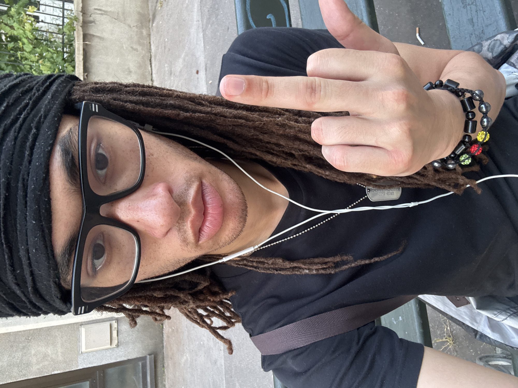 A close-up shot of a person with dreadlocks and glasses is outdoors. The person is wearing a black shirt and a black head covering. They have a bracelet on their wrist, and they are wearing headphones. The person is giving the middle finger. A dog tag hangs from a chain around their neck. The location may be Paris, France.