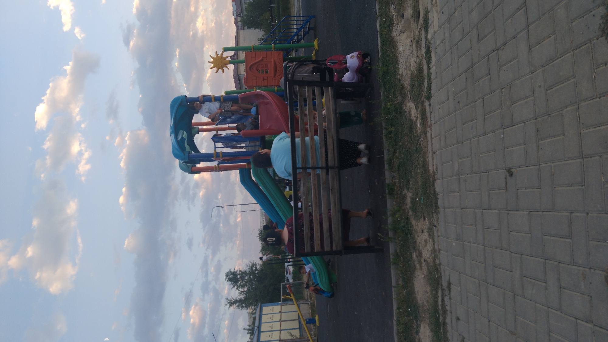 An outdoor, medium shot captures a playground scene at dusk. A large, multi-colored play structure dominates the center, featuring slides of green, blue, and red, along with platforms and climbing elements. Several children are visible on and around the structure; one child stands at the top of the red slide, holding onto the railing. Two women are seated on a wooden bench in the foreground, facing away from the camera, observing the playground. A small pink toy car is parked near the bench. The ground of the playground is black rubberized material, and a paved walkway of grey rectangular bricks runs along the bottom of the frame. In the background, trees and buildings are dimly visible under a sky filled with soft, scattered clouds and the crescent moon.