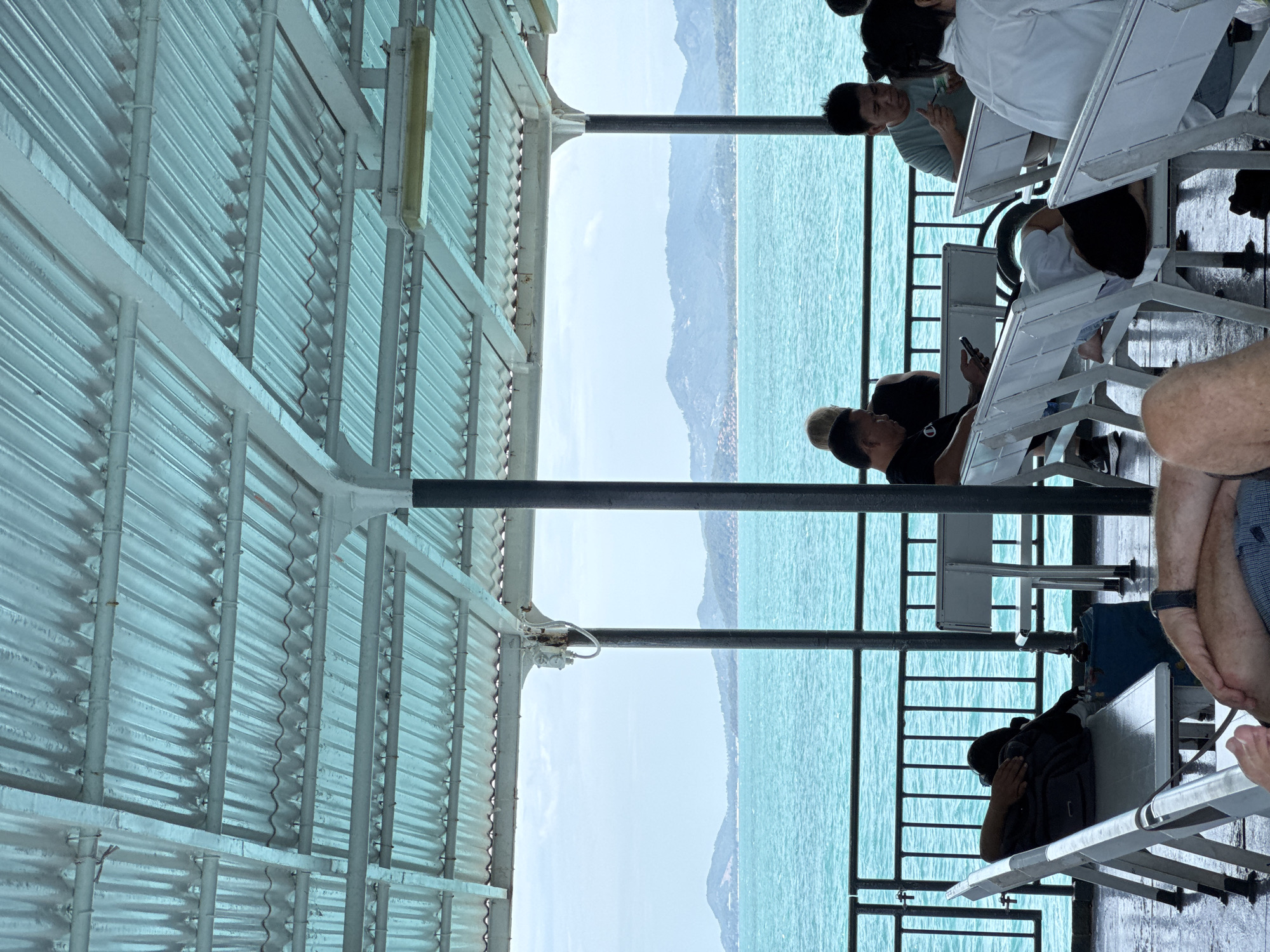 A man is resting on a bench under a corrugated metal roof on a ferry, with his head on a backpack. The ferry is on the water, with a clear view of distant islands and mountains under a cloudy sky. Several other people are seated on benches on the ferry, some looking at their phones or eating. The overall scene suggests a journey across the water. The location is in the city of Unknown, Thailand.