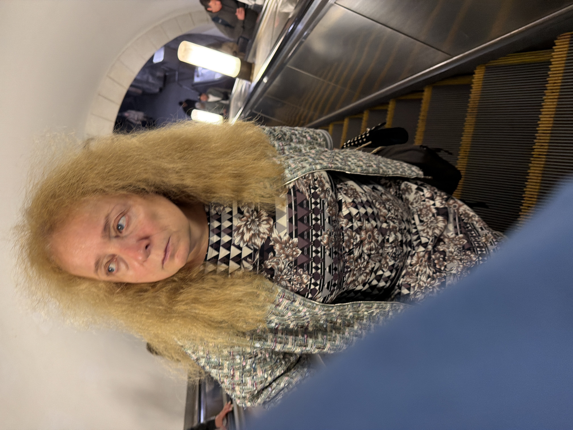 A woman with blonde, frizzy hair stands on an escalator in a Moscow metro station. She wears a patterned jacket over a patterned dress. The escalator has yellow warning strips. In the background, other people are visible, and there are two tall, cylindrical light fixtures. The scene appears to be within the arched structure of the metro station.