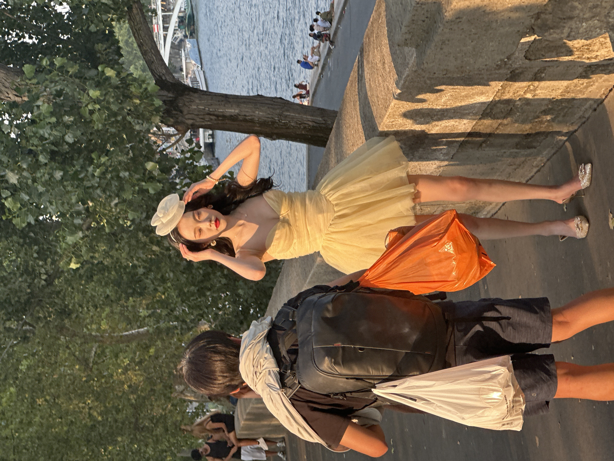 The image shows a woman in a yellow strapless dress and a white headpiece standing on a stone walkway next to a river. She is adjusting her headpiece with her right hand. The woman is wearing high heels and has long dark hair. She is holding an orange plastic bag in her left hand. To her left, a person in a black t-shirt and dark shorts with a backpack is walking away from the camera. This person is carrying a white plastic bag in their right hand. In the background, several people are sitting along the riverbank, and boats are visible on the water. A large tree with green leaves is to the right of the woman. The scene appears to be outdoors during daylight, with warm sunlight casting shadows on the stone walkway. This location is consistent with the Seine River in Paris, France, given the architectural style of the bridge in the background and the presence of boats commonly seen on the Seine.