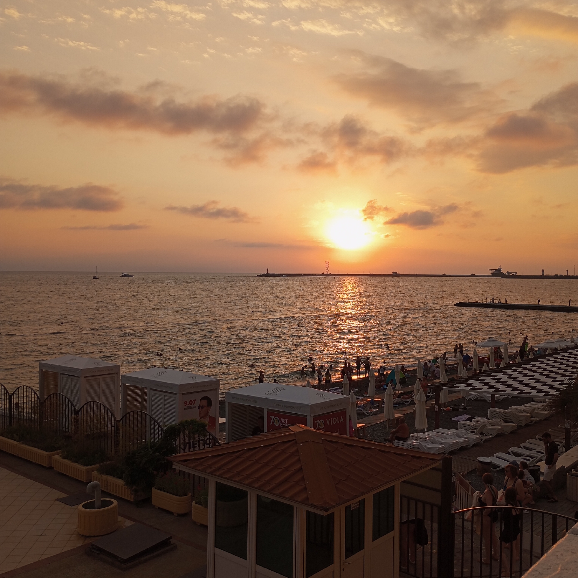 The image depicts a sunset scene at a beach in Sochi, Russia. The sun is low on the horizon, casting a warm, orange glow across the sky and the sea. The water is rippled and reflects the sunlight.
In the foreground, there are several white cabanas or changing rooms lined up along the beach. Some have advertisements on them. There are also rows of empty lounge chairs and umbrellas, suggesting a beach resort setting. People are visible in the water, swimming and wading, and on the beach, walking and sitting. A checkered pattern of black and white umbrellas or awnings is prominent on the right side of the beach.
Further out in the sea, there are a few boats, including a sailboat and a larger motor yacht. A long pier or breakwater extends into the sea, with a lighthouse and some industrial structures visible in the distance. The sky is filled with soft, wispy clouds, tinged with orange and pink from the sunset. The overall atmosphere is peaceful and relaxing, characteristic of a summer evening at the beach.
The location is identified as a beach in Sochi, Russia, likely along the Black Sea coast. The presence of beach amenities, the type of boats, and the general architecture of the cabanas are consistent with tourist destinations in this region.