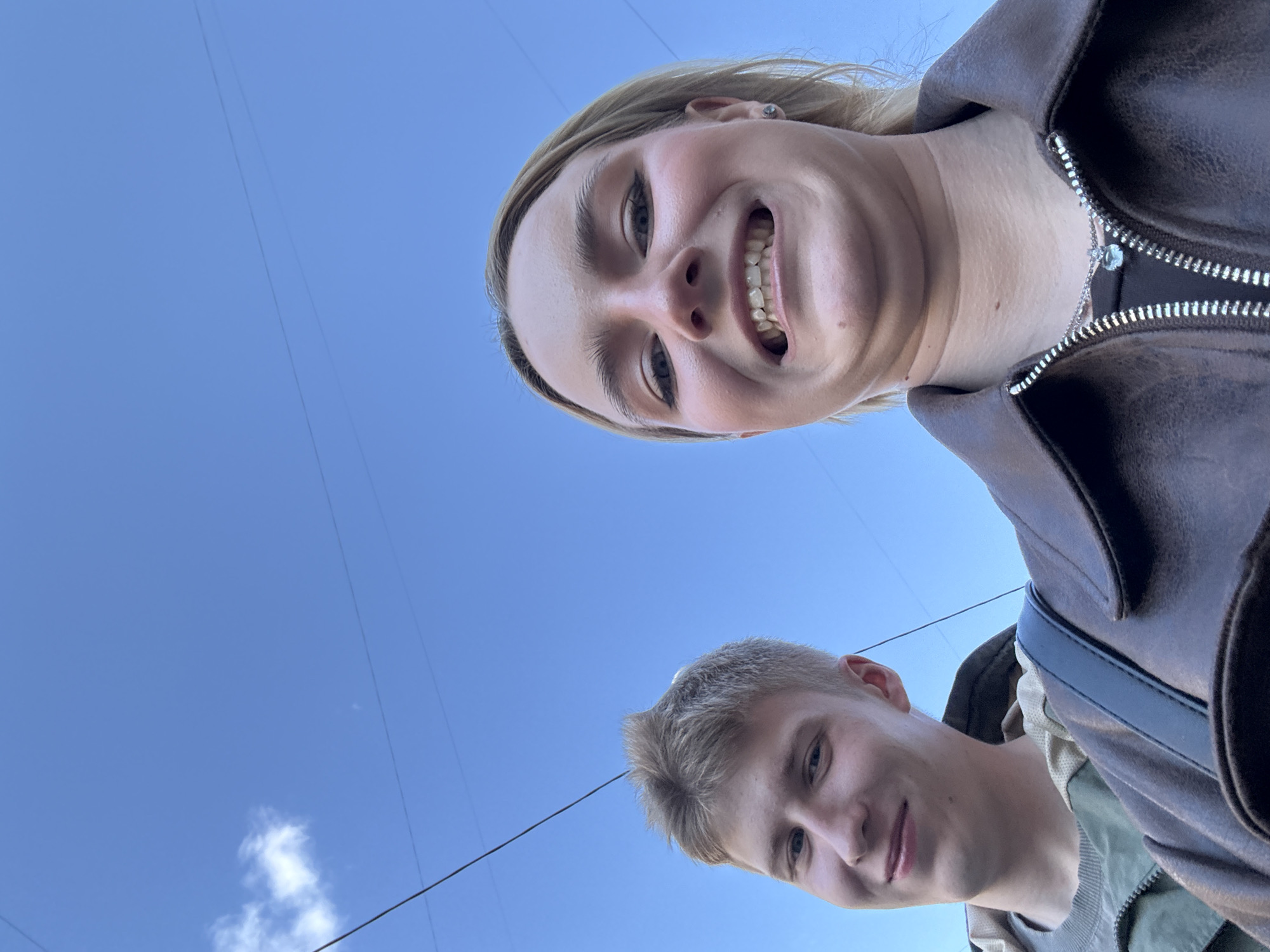 A low-angle shot shows two people against a blue sky with a few clouds. The person in the upper frame is a fair-skinned female with blonde hair, smiling broadly. She is wearing a brown jacket with a zipper and a silver necklace. Below her is a fair-skinned male with short, light-colored hair. He is also smiling. Part of a jacket with a strap is visible on his right. Power lines are visible in the background. The photo was taken in Vsevolozhsk, Russia, as evidenced by the presence of utility infrastructure common to urban and suburban areas in the region.