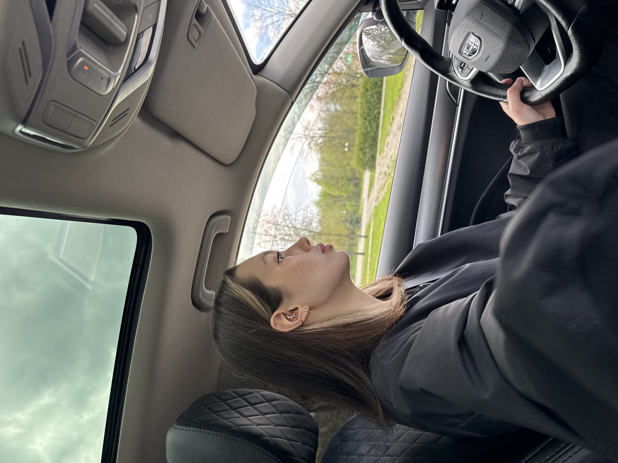 A young woman with long, dark brown hair with lighter highlights is seated inside a car, likely in the driver's seat. She is viewed from a side profile, looking upwards and slightly to the left with a contemplative expression. She wears a dark, possibly black, jacket. Her right hand rests on the steering wheel. Visible on her right ear are multiple piercings, including a prominent curved earring in her upper cartilage and a stud in her lobe connected by a delicate chain.
The car's interior is visible, featuring quilted black seats. The steering wheel has a central emblem that resembles a shield or crest, and the word "AIRBAG" is visible beneath it. An overhead console is present on the car's ceiling, featuring several buttons, one of which has a small orange indicator light. A sun visor is visible on the passenger side.
Through the car's windows, a bright daytime scene unfolds, characterized by a partly cloudy sky and verdant trees with light green foliage, suggesting spring or summer. A patch of what appears to be a paved path or road is also visible in the distance. The overall setting is natural and outdoor, seen from within the vehicle.