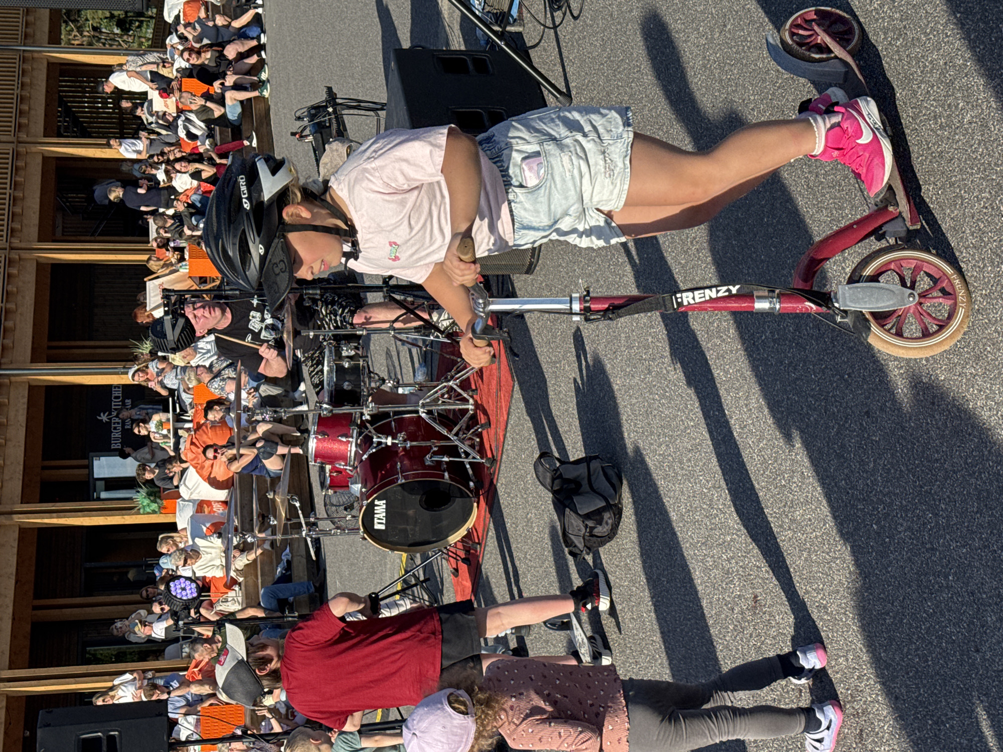 The image depicts an outdoor scene with several people gathered. In the foreground, a person wearing a helmet and a pink t-shirt is standing on a red scooter. They are holding the handlebars and their right foot is on the scooter deck, while their left leg is extended, with a pink sneaker. The scooter has the brand name "FRENZY" visible on its handle.
Behind the person on the scooter, a drum set is set up on a red rug. The drum set appears to be a Tama brand. Several people are seated or standing around the drum set and in the background, some appearing to be an audience or participants in an event. The setting seems to be an outdoor venue with wooden structures and a paved ground.
The overall lighting suggests it is daytime, possibly late afternoon or early evening, given the long shadows cast by the people and objects. The weather appears to be pleasant and sunny. Some visible text includes "FRENZY" on the scooter and "TAMA" on the bass drum. There's also a sign that says "BURGER KITCHEN PAN AS BAR" on a building in the background. The general mood appears to be relaxed and communal, possibly at a small outdoor festival or gathering.