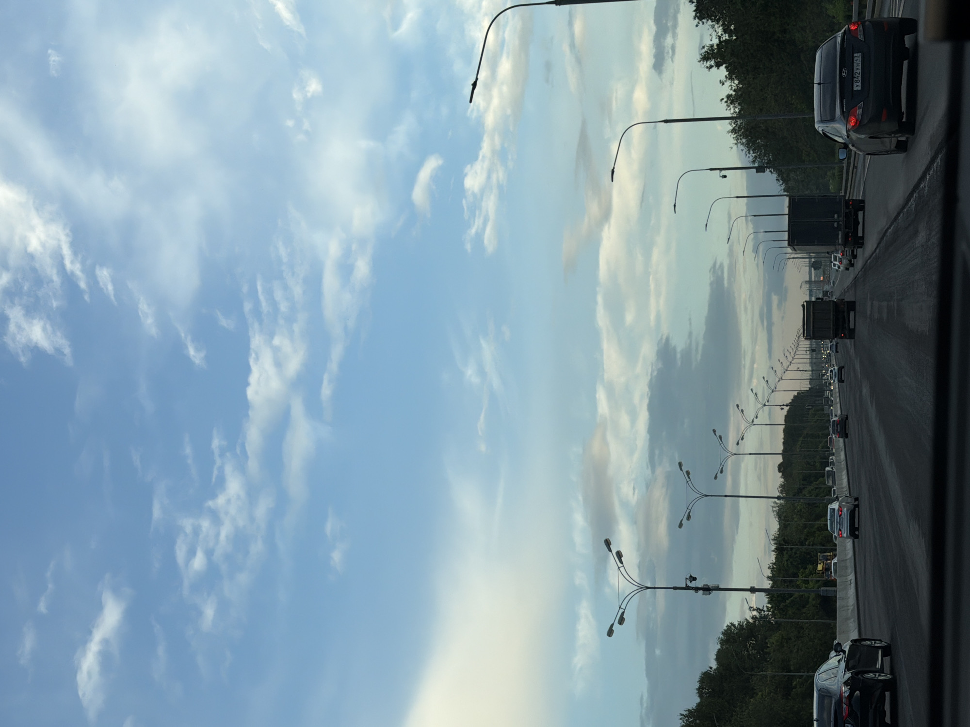 The image depicts a view from a vehicle traveling on a multi-lane highway, likely an overpass, in Moscow, Russia. The sky is partly cloudy with a mix of blue sky and white and gray clouds. A line of streetlights with multiple lamps runs along the right side of the highway, and similar streetlights are visible in the distance. The highway is bordered by trees on both sides, indicating a suburban or exurban setting. Several vehicles, including cars and trucks, are visible on the highway, all traveling in the same direction. One of the cars, a black sedan in the foreground on the right, has a visible license plate with Russian characters and numerals. The overall scene suggests a typical day of traffic on a major roadway in Moscow.