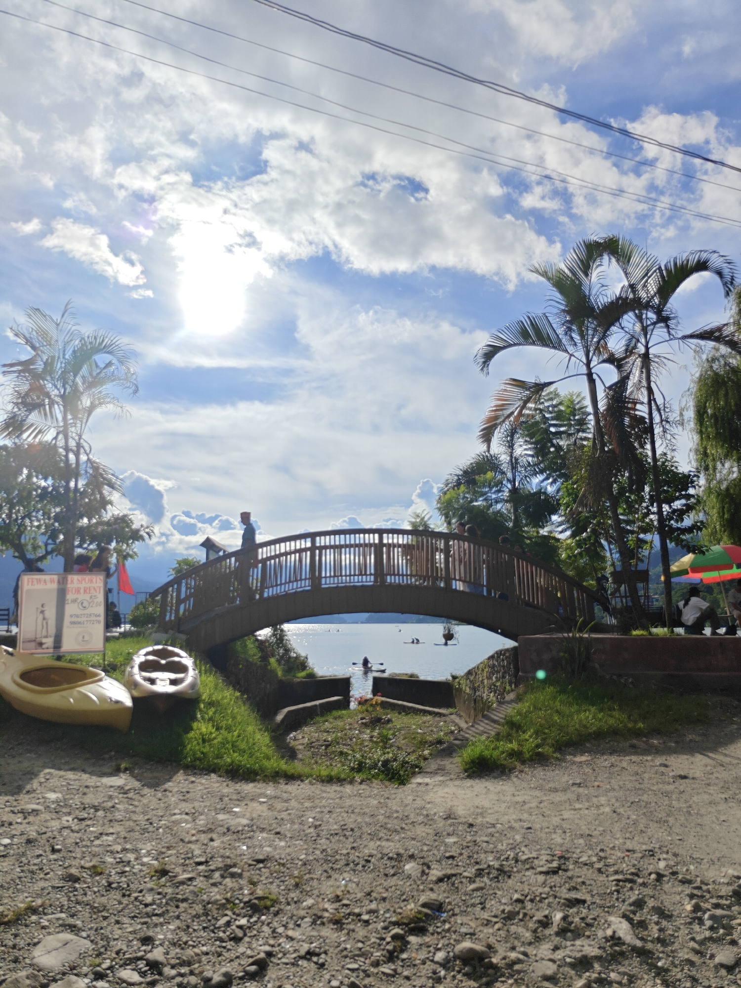 The image captures a scenic view of Phewa Lake in Pokhara, Nepal, under a partly cloudy sky with the sun shining brightly. A wooden bridge arcs gracefully over the water, with several people visible on and near it. On the left side of the foreground, two kayaks are pulled up on the grassy bank. One is a light yellow, and the other is a darker shade, possibly brown or grey. Behind them, a sign reads "FEWA WATER SPORTS FOR RENT". Palm trees and other lush greenery surround the area, adding to the tropical ambiance. In the distance, the calm waters of Phewa Lake reflect the sky, and a few small boats or kayaks are present on the water. The overall scene suggests a peaceful and recreational setting.