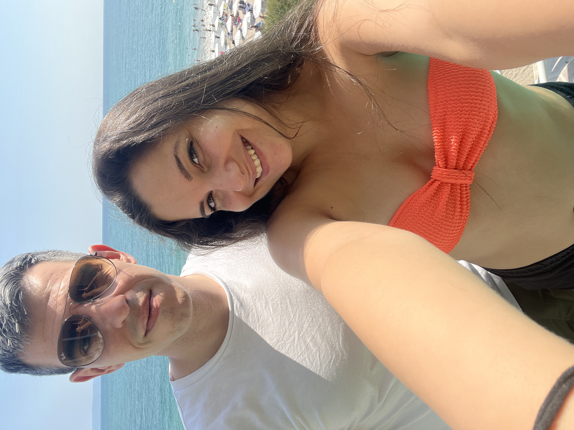 A close-up selfie shows a man and a woman at the beach in Pomorie, Bulgaria. The man, wearing sunglasses and a white t-shirt, is on the left. The woman, smiling and wearing an orange bikini top, is on the right. The background shows the Black Sea, a beach with umbrellas and people, and a clear blue sky.
