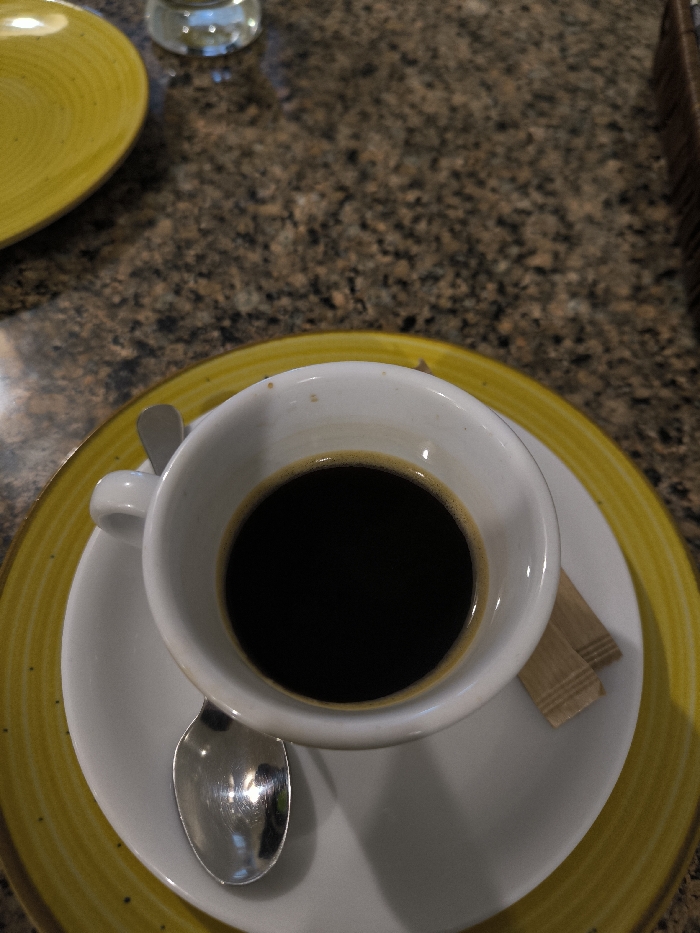 The image displays a high-angle view of a single serving of black coffee on a table. The central subject is a white ceramic cup, approximately three-quarters full of a dark brown liquid, consistent with black coffee, topped with a faint light brown crema ring. The cup rests on a white ceramic saucer, which in turn is placed on a larger yellow ceramic plate featuring a textured, ridged rim. A small, polished silver-colored spoon lies on the white saucer to the lower-left of the cup. To the lower-right of the cup on the saucer, two small, rectangular, light brown paper packets, likely containing sugar or sweetener, are present.
The table surface beneath these items is dark brown with irregular lighter and darker speckles, characteristic of granite or a similar stone material. In the upper-left quadrant of the frame, a partial view of another yellow ceramic plate, matching the one under the coffee, is visible. Above this plate, a blurry, transparent glass object, possibly a water glass, is partially discernable. A small portion of a brown, woven object, possibly a basket or container, is visible along the right edge of the frame in the upper section. No people are visible.
The specific location within the city of Vinnytsia, Ukraine, cannot be determined from the visual information present in this image.