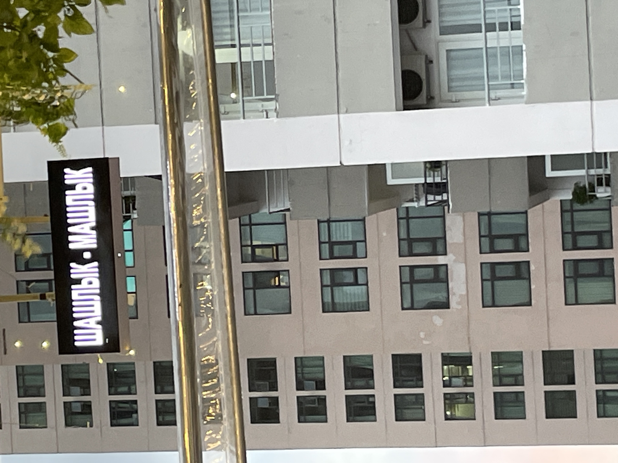 A slightly rotated medium shot shows a building in Bac Nha Trang Ward, Vietnam. In the foreground, a tall, shiny metal pole stands slightly off-center. To the left of the pole, a vertical rectangular sign reads "SHAШLYK - МАШЛЫК" in bright white Cyrillic letters against a black background. Behind the sign and pole, a multi-story building dominates the scene. The building's facade is light gray with numerous windows arranged in neat rows and columns. Some windows have dark frames, while others are lighter. Air conditioning units are visible near the top of the building. The sky is not visible. A few green leaves are visible in the upper left corner.