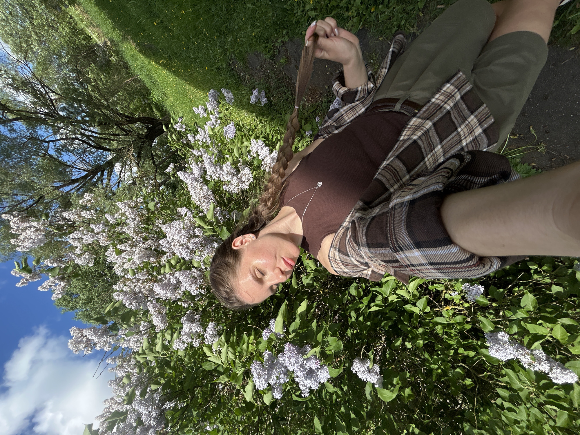 A woman with light skin and brown braided hair is lying amidst a bush of lilac flowers in the city of Борщевка Малая, Russia. She is wearing a brown tank top, olive green pants, and a brown and beige plaid shirt. The woman is holding the end of her braid in her right hand. In the background are a tree, grass, and a path. The sky with white clouds is visible in the upper left corner.