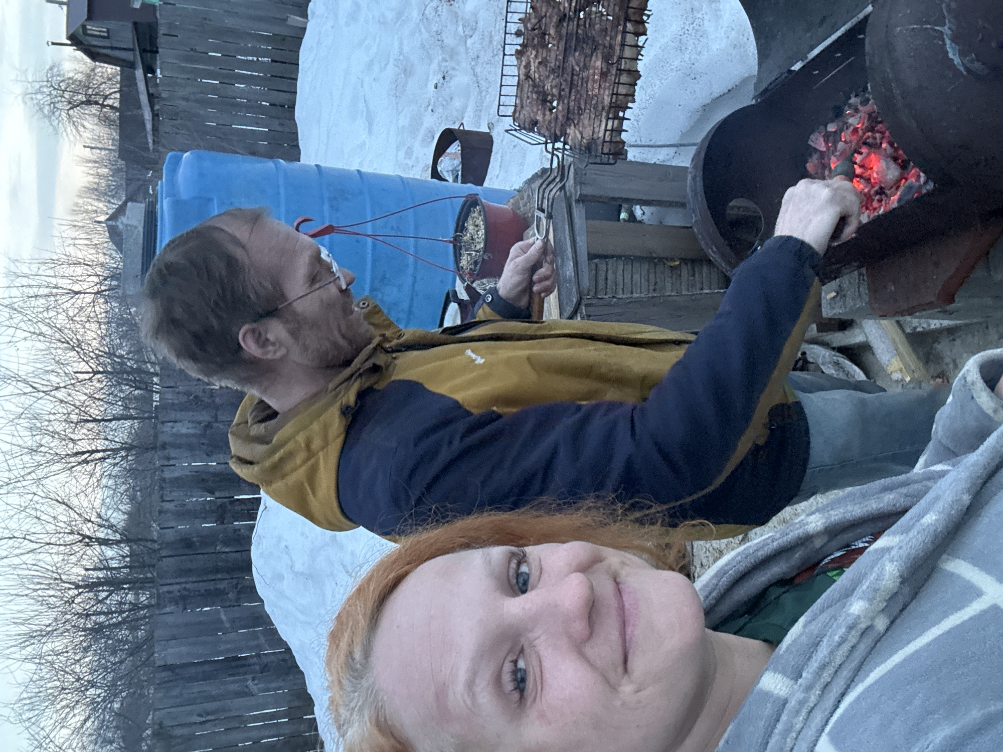The image depicts an outdoor scene in a residential backyard or dacha plot within the city of Chaikovsky, Russia, characterized by snow-covered ground and bare trees. Two individuals are present.
In the foreground and to the left, a female subject with reddish-blonde hair, fair complexion, and light eyes is positioned, smiling directly towards the camera, suggesting she is taking a selfie. She is wrapped in a grey and white patterned blanket or robe, over a green garment.
In the middle ground, to the right of the female, a male subject with short brown hair, a receding hairline, and eyeglasses is observed. He is wearing a yellow and dark blue two-tone jacket and blue jeans. He is actively tending a dark-colored, barrel-style metal grill containing glowing red charcoal. His right hand holds metal tongs, while his left hand is near a rectangular metal grilling basket, which holds multiple cuts of meat (likely shashlik), positioned directly over the coals. The meat appears partially cooked.
Behind the male, a large, bright blue plastic barrel stands upright. A smaller red pot, suspended by red cords, hangs near his head. A wooden plank fence forms the immediate background. Beyond the fence, significant patches of snow are visible on the ground. Several bare-branched trees are scattered across the distant background, with parts of residential structures, including a house with a dark roof, also discernible. The sky is overcast, indicating dim, natural light, likely late afternoon or early evening, consistent with the snowy, cold environment.