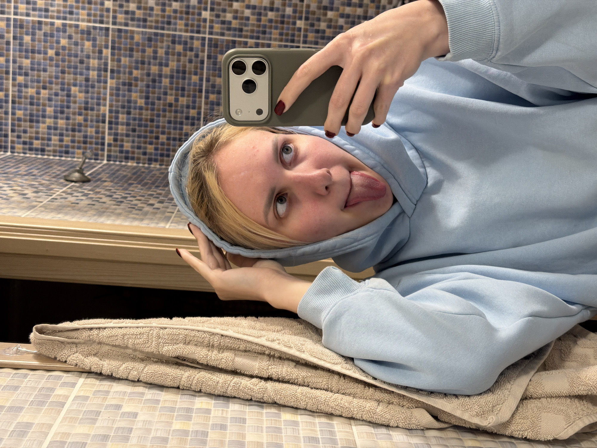 The image shows a young woman with blonde hair wearing a light blue hoodie. She is holding a dark green smartphone in her right hand, taking a selfie. Her tongue is sticking out, and her left hand is touching the hood of her sweatshirt, resting on her head. She is looking to her right. In the background, there is a towel hanging on a hook on a tiled wall, and a doorway. The tiled wall has a pattern of small, square tiles in shades of blue and brown. This appears to be a bathroom or a similar domestic setting. There is no information in the image that indicates the location in the city of Cheltuitori, Moldova.