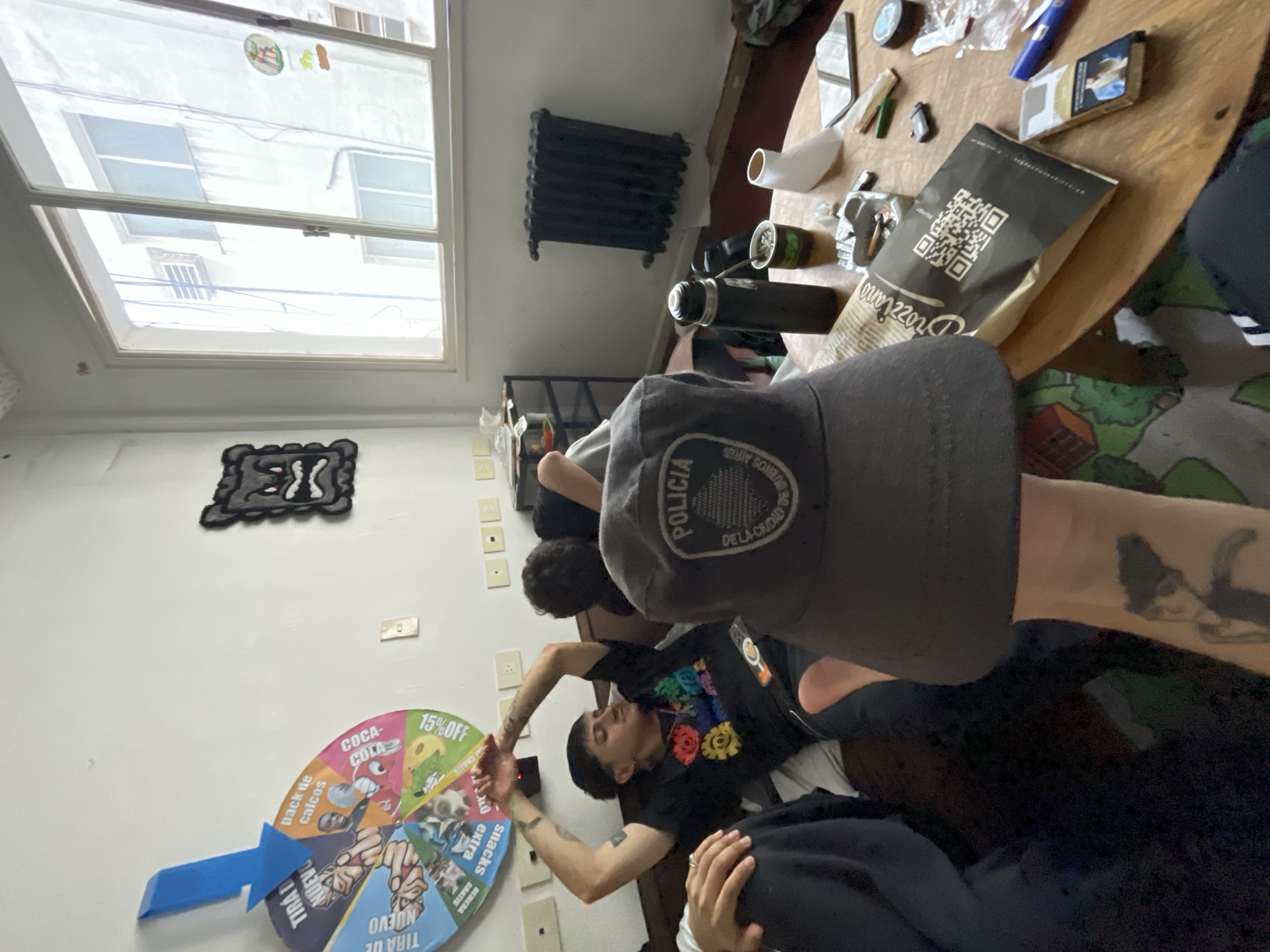 The image captures an indoor scene in Buenos Aires, Argentina, featuring several individuals engaged in a casual gathering during the daytime. The bright light streaming through a large window, which reveals the exterior of another building, indicates the time of day.
In the immediate foreground, a person's arm and upper torso are visible, wearing a dark gray baseball cap. The cap prominently displays the text "POLICIA DE LA CIUDAD DE BUENOS AIRES," clearly identifying the location. The arm has visible tattoos.
Towards the center, another person is lying down, wearing a black t-shirt adorned with a colorful, gear-like or floral graphic design. This individual has their arms slightly raised. Other figures are partially seen, suggesting multiple people are present.
The room features a classic black radiator mounted on the wall below the window. To the right, a round wooden table holds several items, including a black thermos, a traditional mate cup (a cultural staple in Argentina), a cigarette pack, a lighter, and a paper bag marked with a QR code and the brand name "Drozziano."
A colorful spinning wheel game is mounted on the wall to the left, featuring a blue arrow and various prize segments with Spanish text. These include "COCA-COLA," "15%OFF," "pack de calcos" (sticker pack), "snacks extra," "REMERA GRATIS" (free t-shirt), and "TIRA DE NUEVO" (spin again). Above the wheel, a dark, stylized wall decoration is visible. The overall ambiance is relaxed and informal.