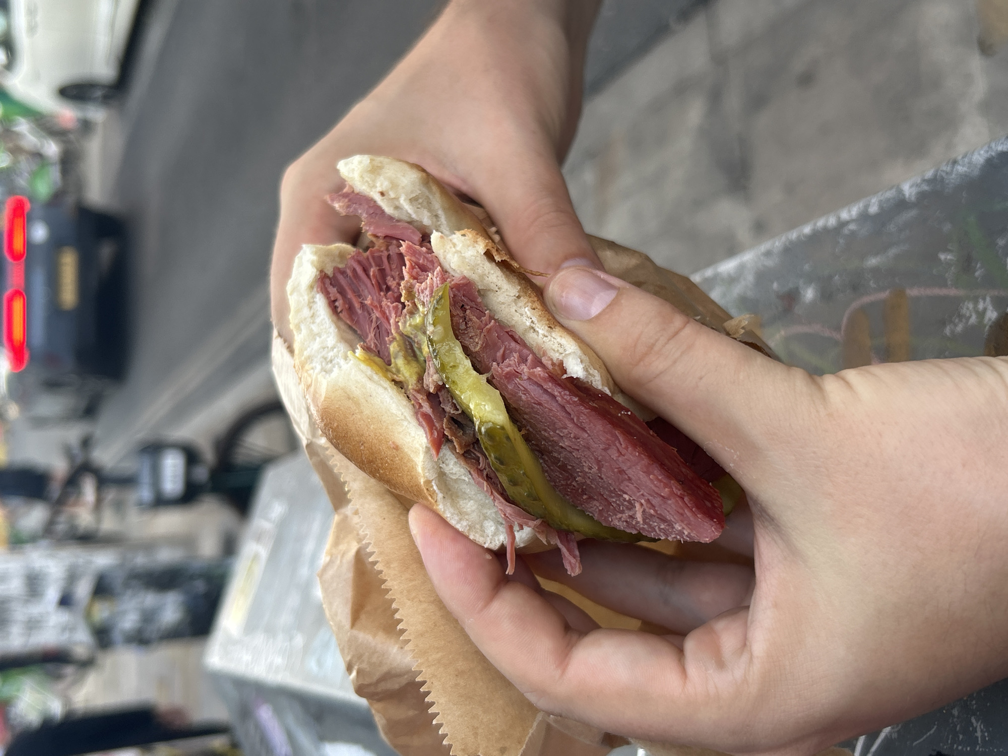A close-up shot shows a person holding a large sandwich filled with thinly sliced meat and pickles. The sandwich is wrapped in brown paper. In the background, blurred out, are cars on a street, suggesting an urban environment. The scene appears to be outdoors, with elements of a city street visible. The overall impression is of a casual, on-the-go meal.