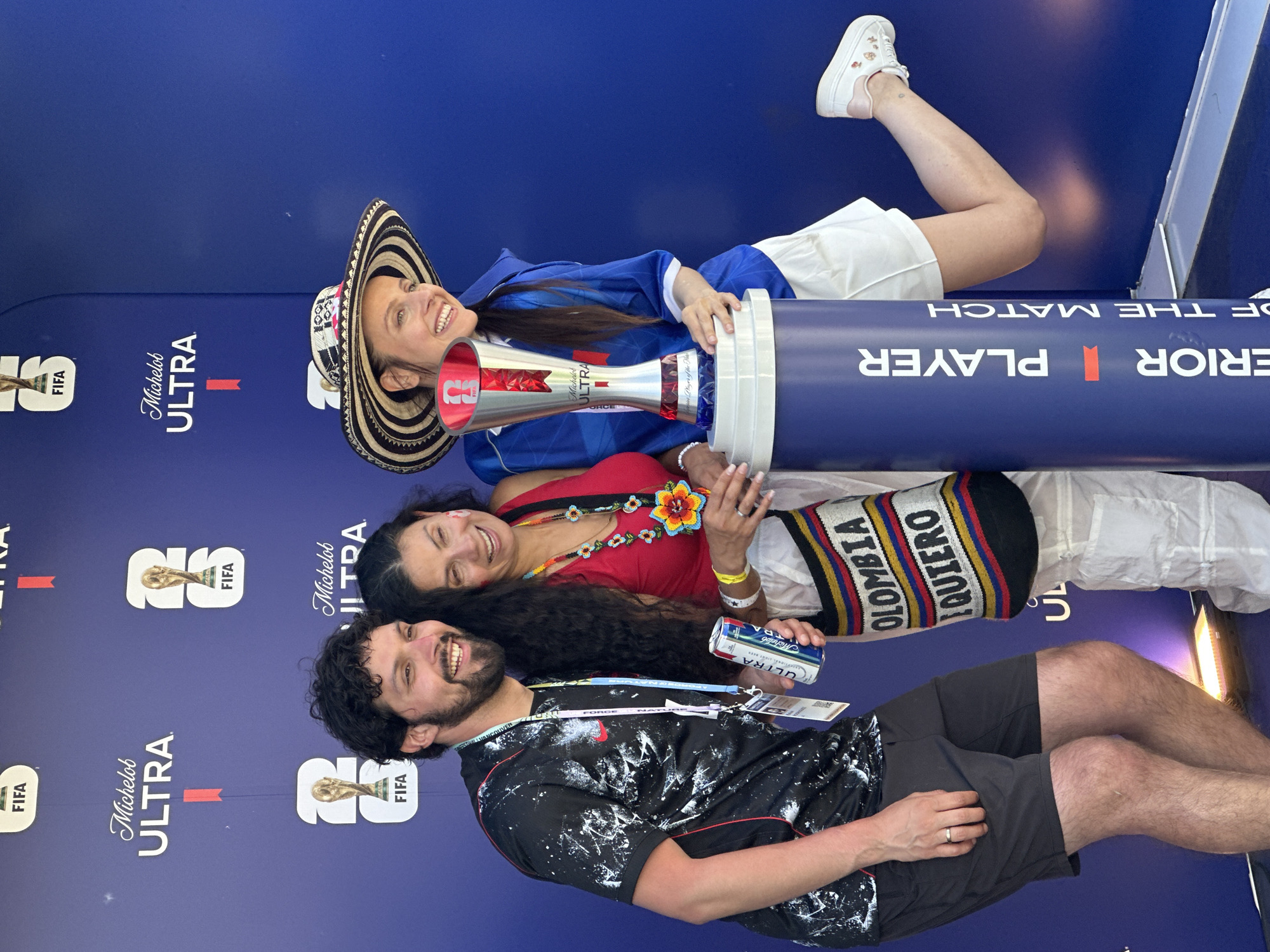 Three individuals are posing for a photo in front of a dark blue backdrop with the Michelob Ultra and FIFA logos. On the left, a man with dark curly hair and a beard is wearing a black t-shirt with white paint splatters and black shorts. He is holding a can of Michelob Ultra in his right hand. In the center, a woman with long dark hair is wearing a red sleeveless top and white pants. She has a colorful beaded necklace and a bag slung over her shoulder with the word "COLOMBIA" visible. On the right, a woman wearing a wide-brimmed black and white striped hat, a blue shirt, and white shorts is leaning on a trophy on a cylindrical stand. The trophy is silver and red, with "Michelob ULTRA" and "FIFA" branding. The cylindrical stand has "PLAYER OF THE MATCH" printed vertically on it. The backdrop has repeated FIFA World Cup trophy imagery. The lighting suggests an outdoor event, likely during the daytime. The presence of the FIFA World Cup trophy and associated branding suggests this is likely a promotional event related to the tournament. While the backdrop doesn't explicitly mention Vancouver, Canada, such promotional activations for major sporting events are common in cities hosting related fan zones or official viewing parties.