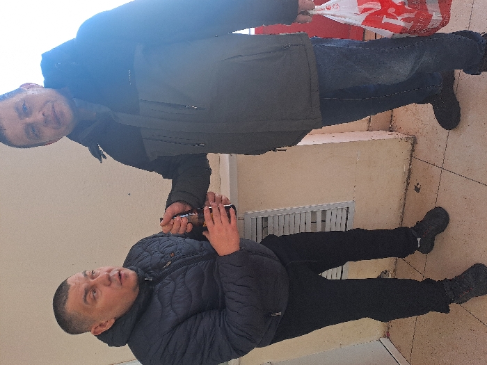 Two adult males are depicted standing on a tiled outdoor surface in Bolshaya Elhovka, Russia. The male on the left, wearing a dark blue puffer jacket, black pants, and dark athletic shoes, holds a dark glass bottle with both hands and appears to be speaking, looking towards the right. The male on the right, dressed in a dark olive green and black winter jacket, blue jeans, and dark shoes, is holding the same dark glass bottle with his left hand, interacting with the first male. He also carries a red and white plastic shopping bag in his right hand. The background features a plain, light-colored wall with a lower ledge and a white horizontal slatted structure. The floor consists of light-colored square tiles.