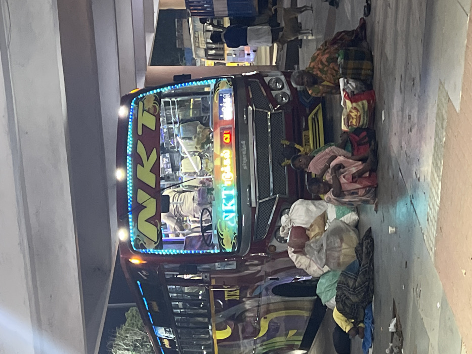The image, when viewed upright, depicts a nighttime scene at what appears to be a bus stop or roadside in Palani, India. A large, maroon-colored bus, heavily decorated with colorful designs and blue LED lights lining its front windshield, is the central subject.
Prominently displayed in yellow letters on the bus's front is "N K T". Below this, in Tamil script, are the characters "என். கே. டி" (N. K. T.) followed by "திருச்சி" (Tiruchy or Trichy) and the number "31", likely indicating the bus company, route, or destination. A four-digit number, "7007", is also visible on a yellow panel near the bus's front. The bus's interior is lit, showing seats and possibly passengers.
Several people are gathered on the paved ground beside the bus. An older woman in a patterned saree sits near the bus's front, with her bags beside her. Further along the side of the bus, three other individuals, possibly women or children, are seated on the ground, surrounded by bundles of belongings, suggesting they are travelers. One person is seen sleeping on the ground, covered by a blanket. In the background, other figures are visible, including a person in white traditional attire and two dogs.
The setting is under an elevated concrete structure, possibly an overpass or building overhang, providing a sheltered area. The artificial lighting, including the bus's illuminated interior and exterior lights, confirms the time of day as night or very early morning. The scene conveys a sense of waiting or rest during a journey.