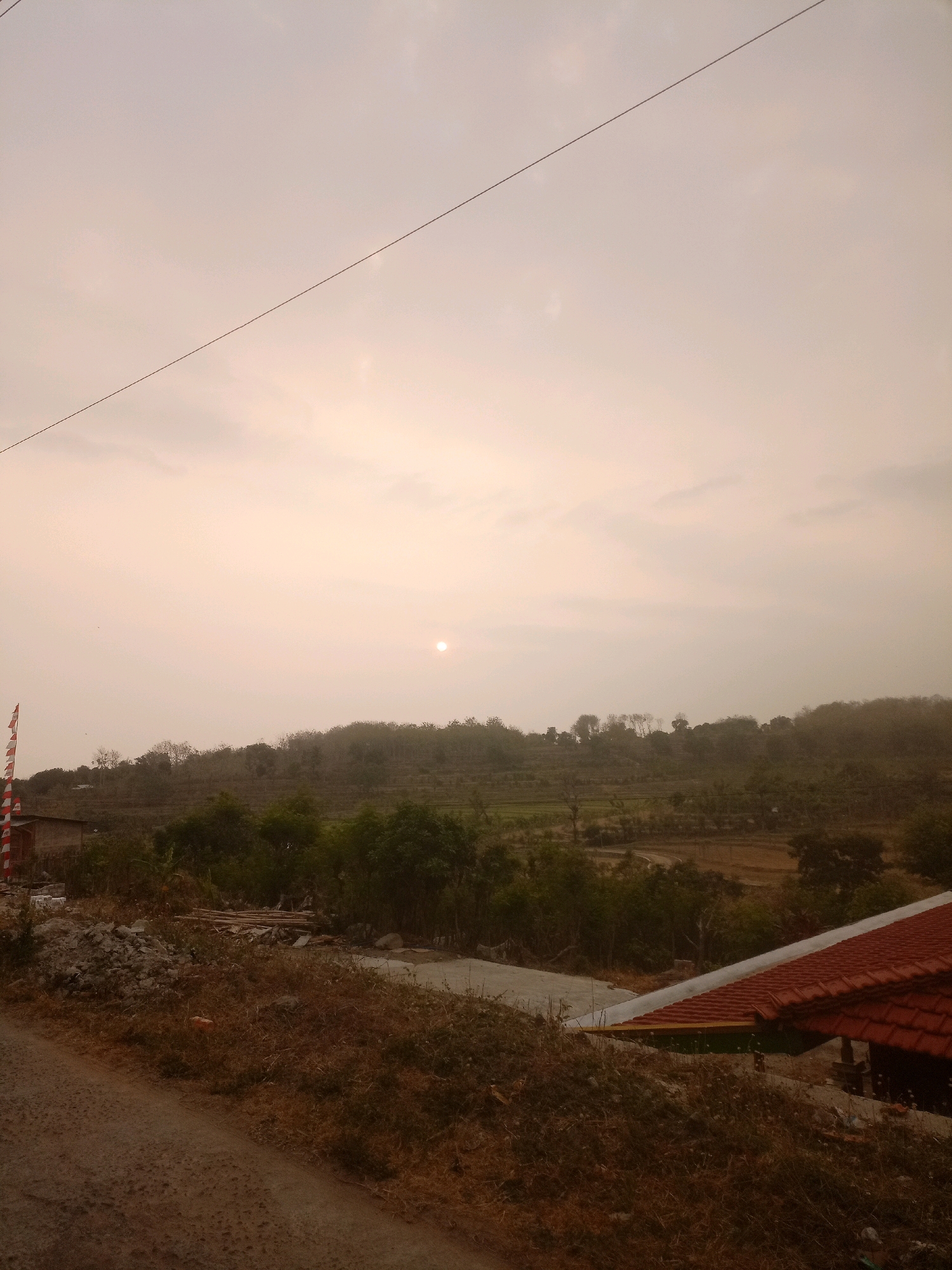 The image shows a rural landscape at dusk. The sky is overcast with a pale, hazy sun visible near the horizon. A thin wire stretches diagonally across the upper portion of the frame. In the foreground, a dirt road curves to the left, bordered by dry, overgrown vegetation. To the right, a house with a red tiled roof is partially visible. Further in the distance, rolling hills are covered with trees and some evidence of terraced fields. A small building and a flag with red and white stripes are visible on the far left of the frame. The overall impression is one of a quiet, natural setting.