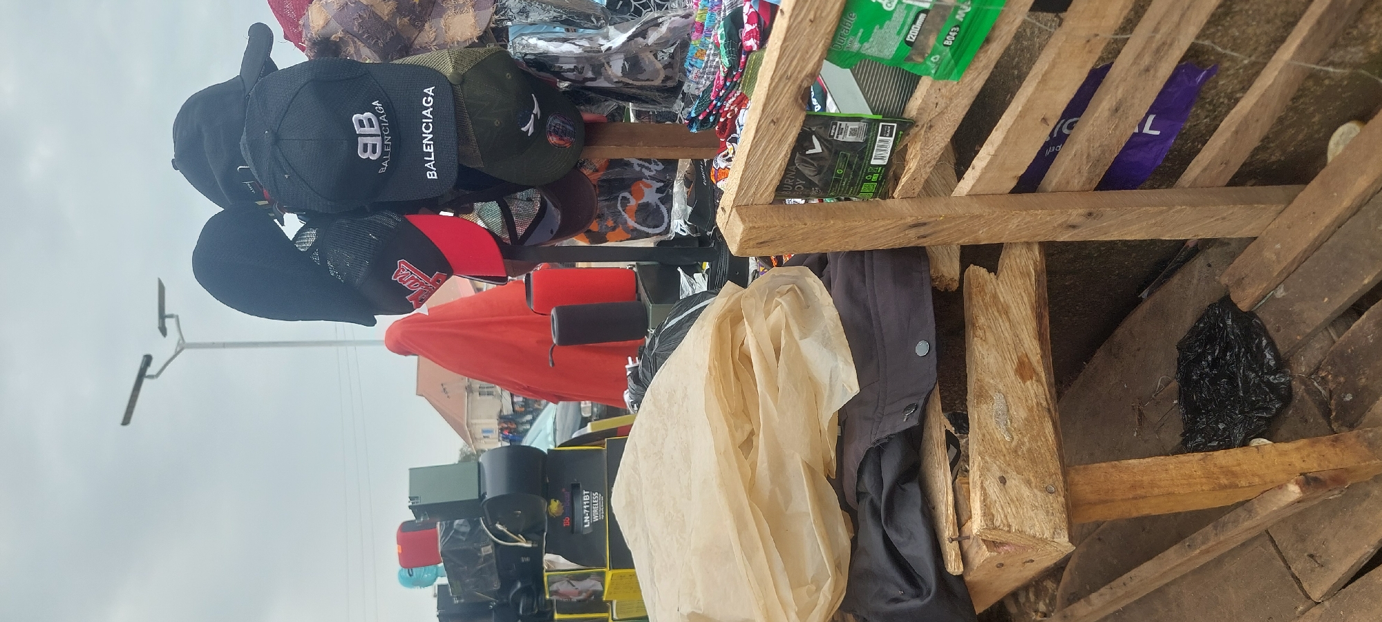 A street market stall is depicted, with various goods for sale under an overcast sky. In the foreground, a wooden pallet supports a stack of clothing and paper-like material, with a black plastic bag on the wooden planks below. The stall displays an assortment of caps, including two with "BALENCIAGA" branding, a red and black cap with "LA" lettering, and a green cap. Behind the caps, a person draped in an orange garment is visible, along with stacked boxes, some labeled "LN-711BT WIRELESS," and cylindrical objects, one red and one black, likely speakers. A lamppost with two solar lights stands in the background. The environment suggests an outdoor commercial setting.