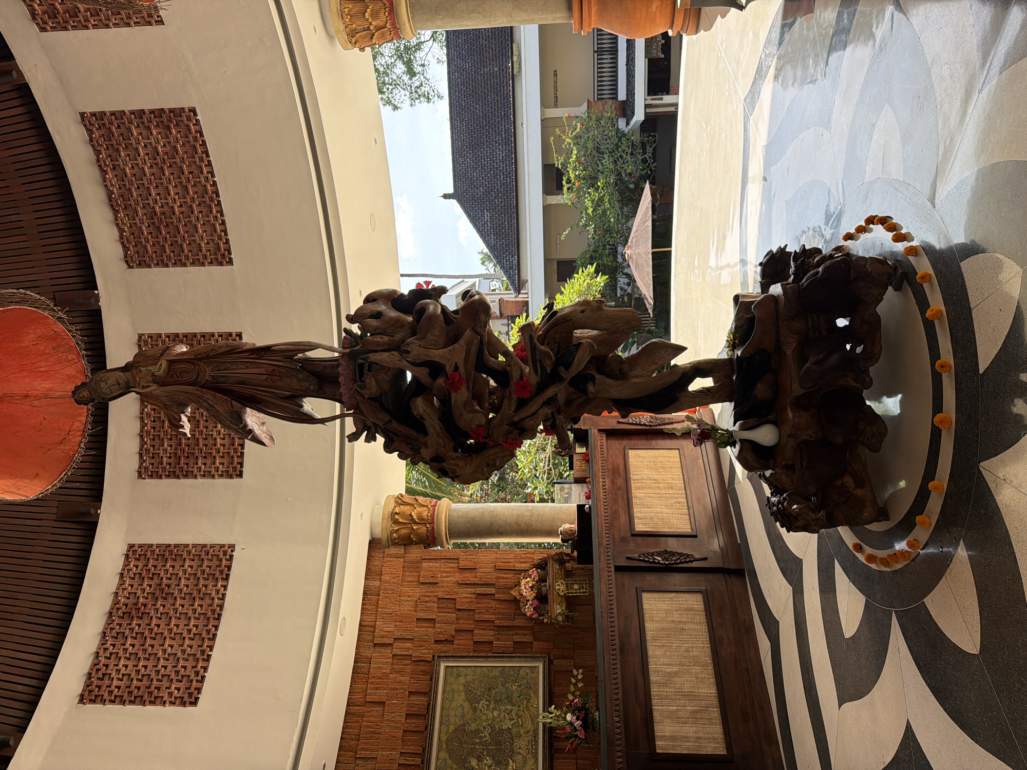 The image captures an interior space in Peliatan, Indonesia, showcasing a wooden sculpture as the focal point. The sculpture is a complex arrangement of carved wood pieces, interspersed with small red flowers, and is situated on a patterned black and white floor. The surrounding architecture includes brickwork walls, decorative columns, and traditional Indonesian design elements. A doorway with wooden panels is visible in the background, along with an exterior view of a building, trees, and a glimpse of the sky. The space is well-lit, possibly from both natural and artificial light sources.